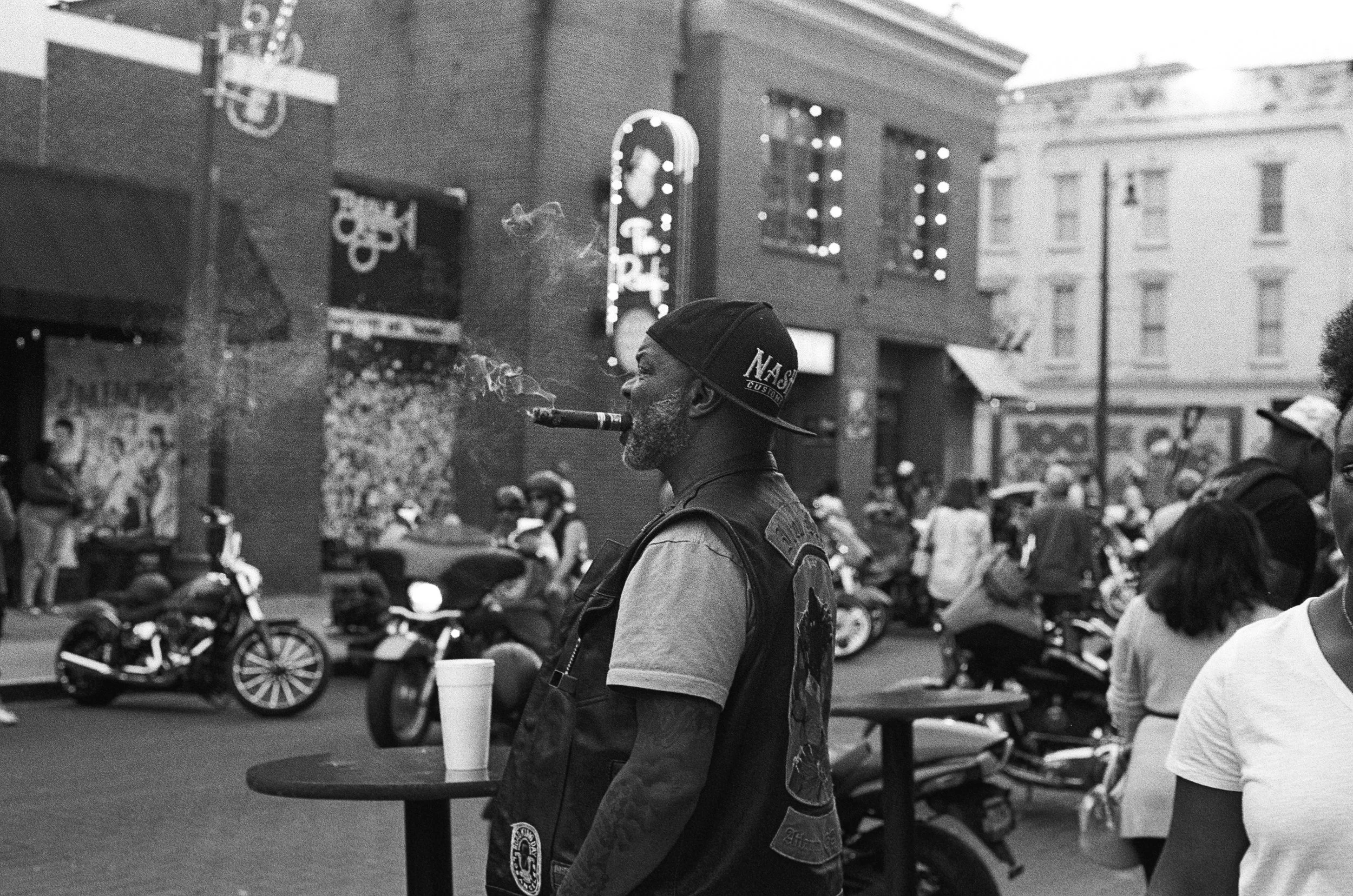 A person wearing a baseball cap with 'Nash' on it, smoking a cigar, at a busy outdoor event with motorcycles and people in the background.