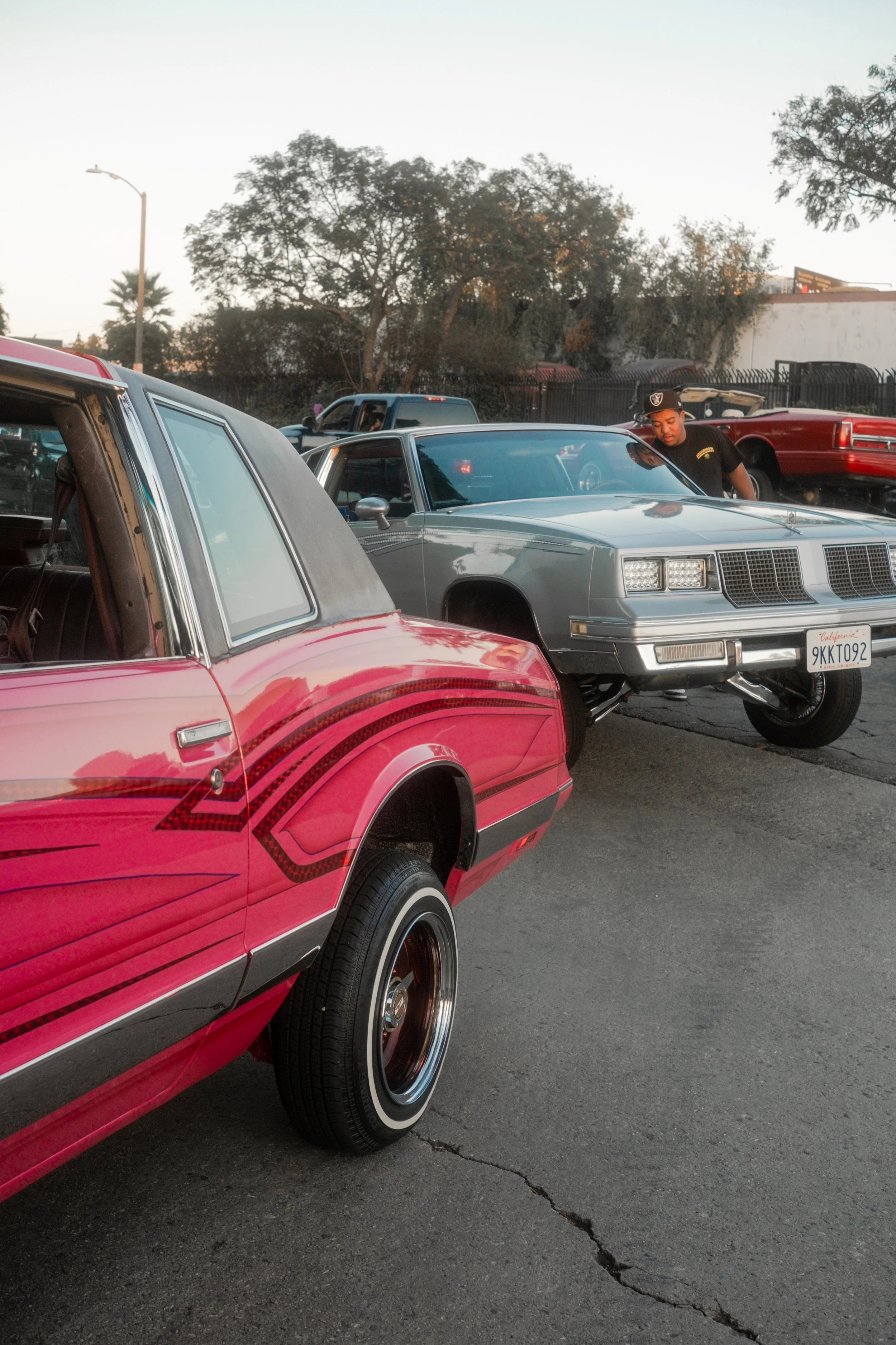 A person looking at a silver vintage car parked in a lot, with other cars and trees in the background during late afternoon or early evening.