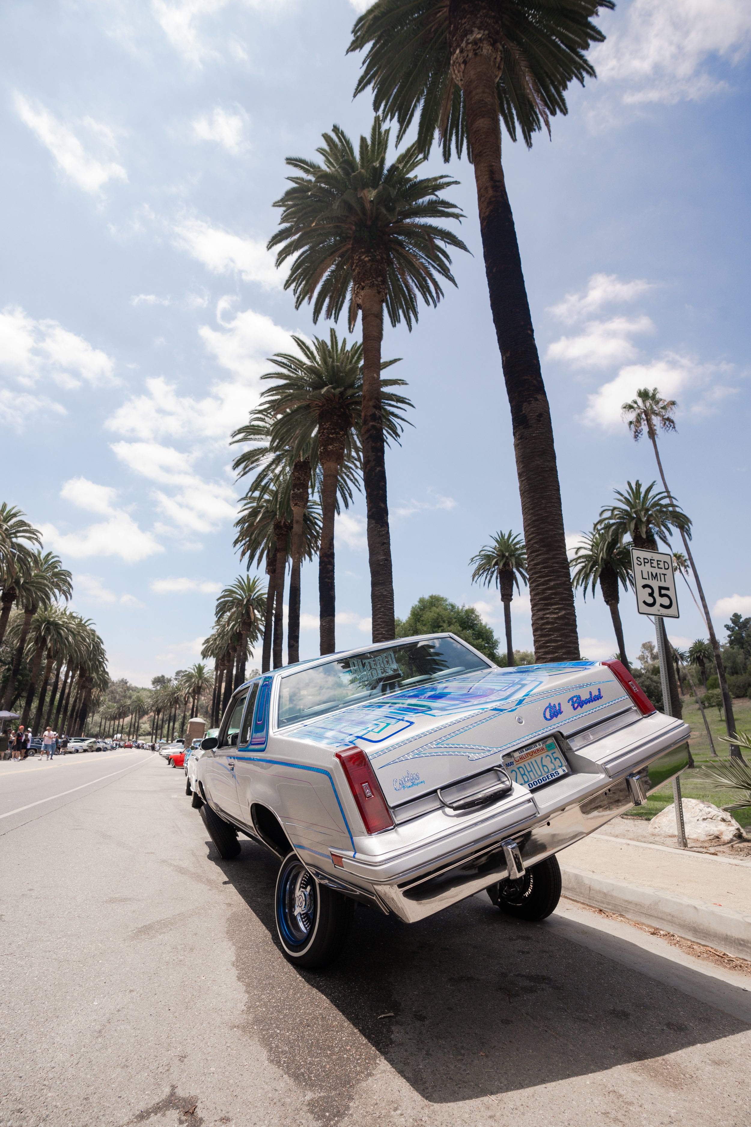 A vintage Cadillac with a custom paint job, tilted on its side in an outdoor parking lot lined with tall palm trees under a partly cloudy sky.