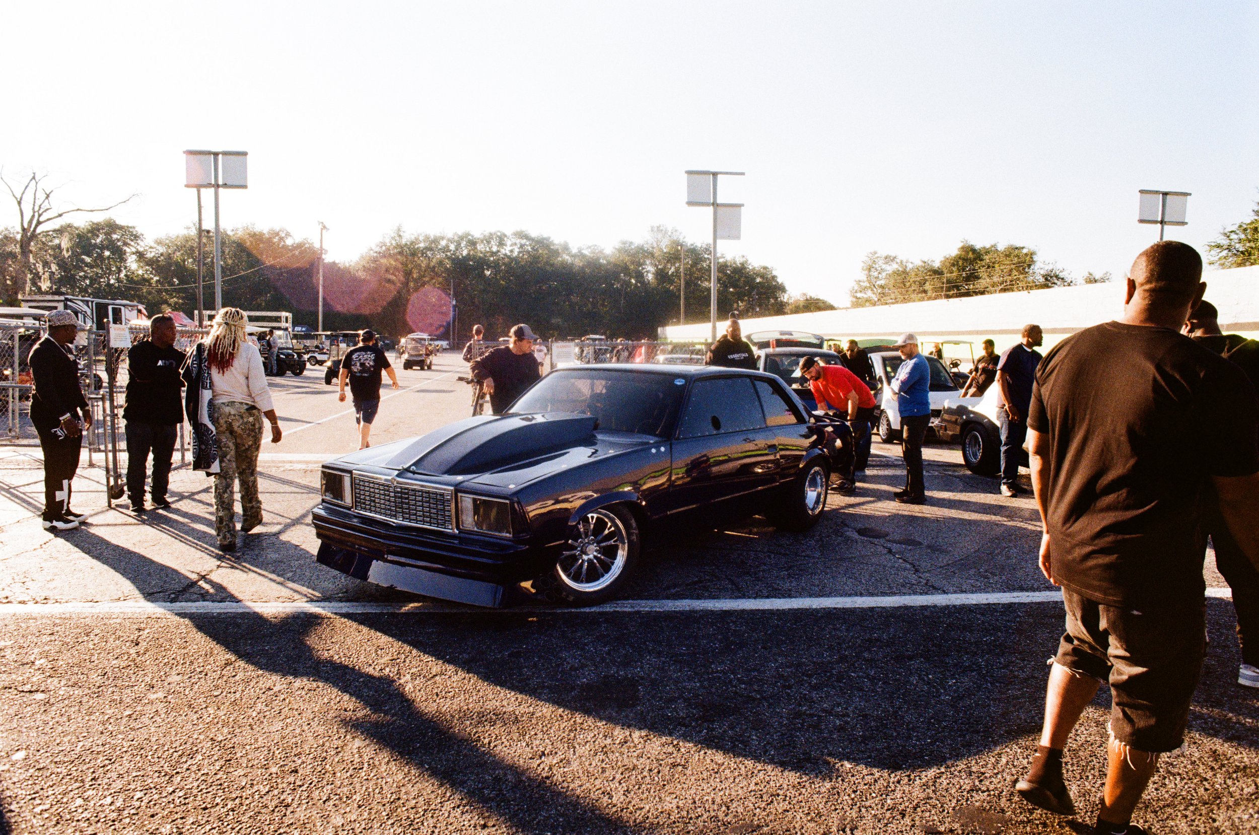 People gathered outside in a parking lot near a display of classic and vintage cars during what appears to be a car show, with sunlight casting long shadows.