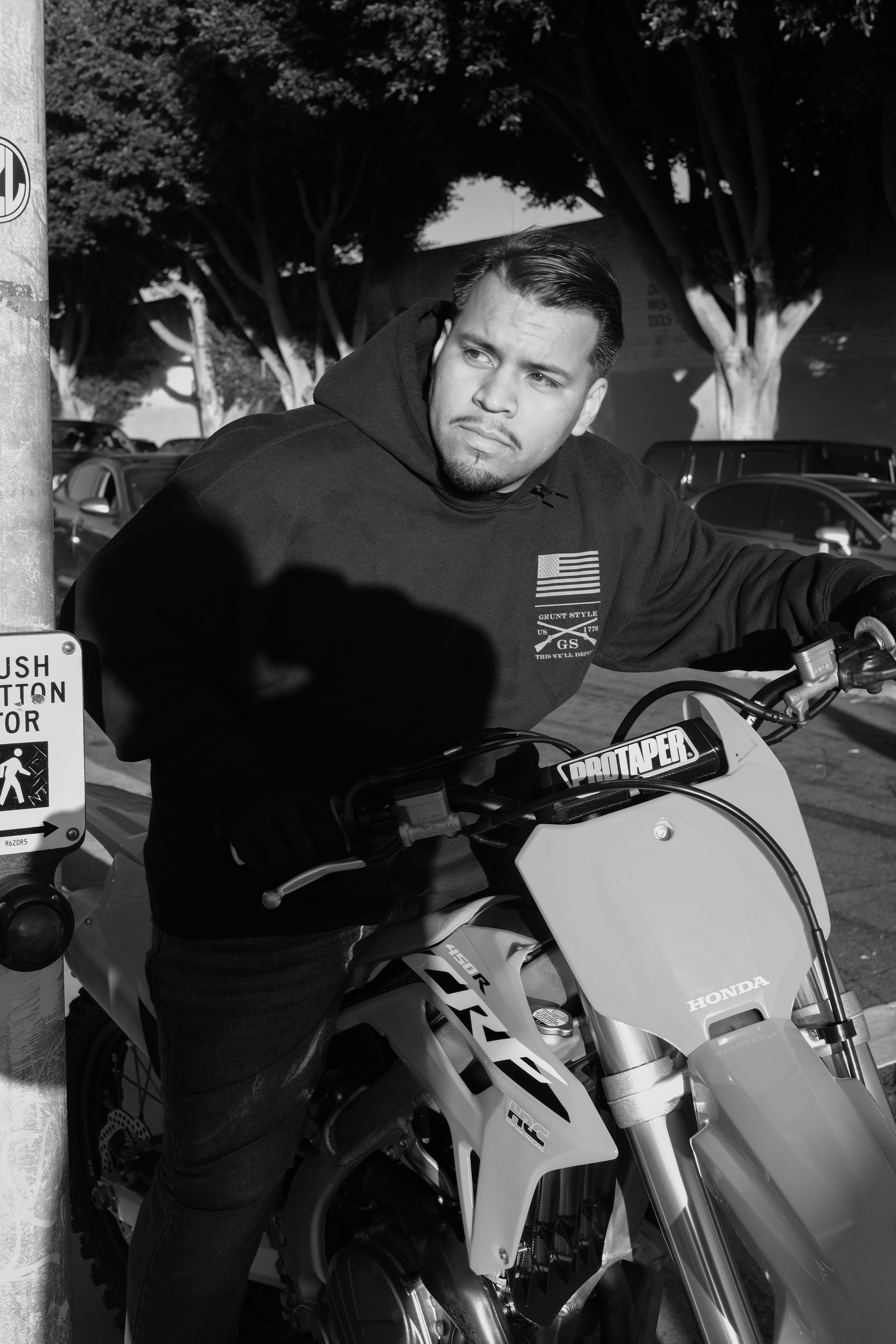 A man with short dark hair and a beard sitting on a Honda dirt bike, wearing a black hoodie with an American flag patch on the chest, in a parking lot with trees and cars in the background. The scene is in black and white.