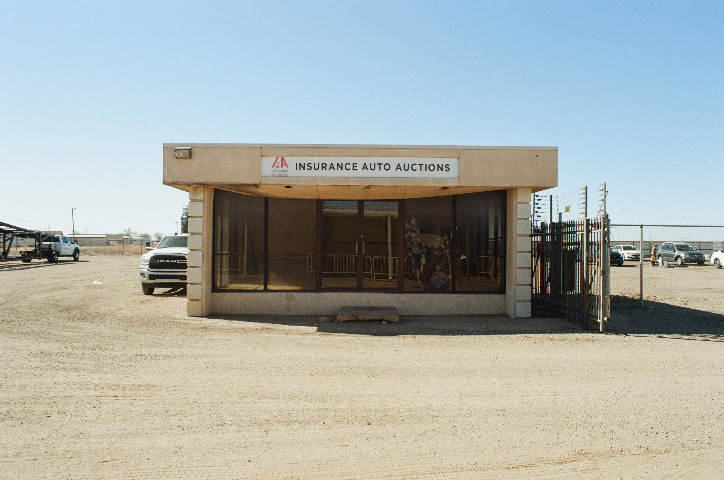 A small beige building with a sign that reads 'Insurance Auto Auctions' and an AA logo. The building has glass doors and is situated in a dusty lot with several parked vehicles and a fence in the background, under a clear blue sky.