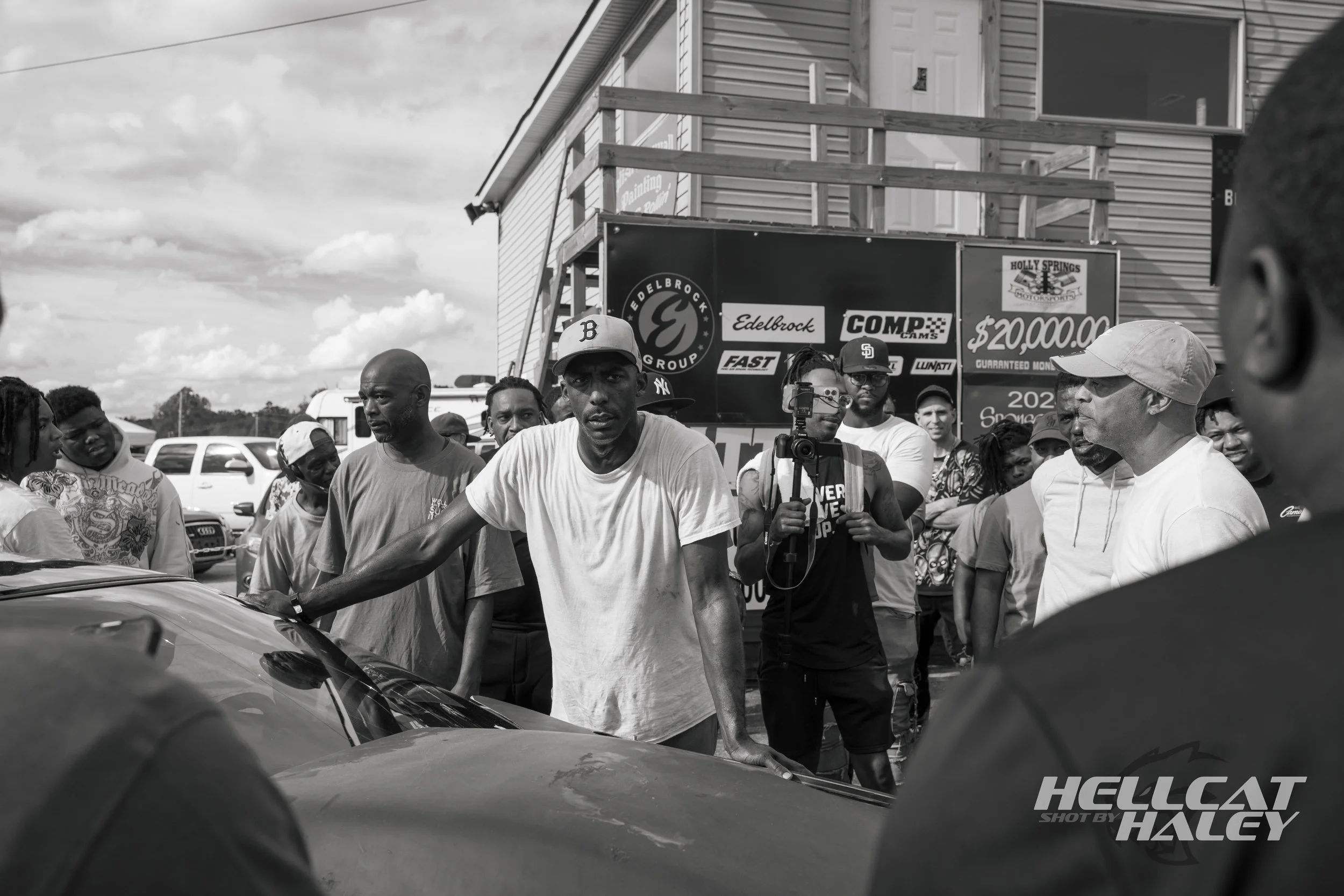 A group of people gathered in a parking lot around a car, with a man wearing a baseball cap and white t-shirt in focus. Behind them is a signboard with logos of racing sponsors and a prize amount of $20,000. The scene appears to be at a racing or car