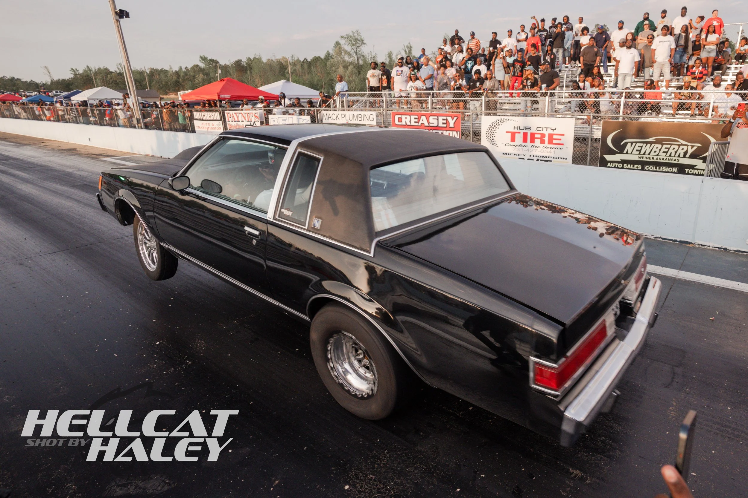 A black vintage muscle car with a black roof is racing on a drag strip with a large crowd of spectators watching from the grandstands. The sky is partly cloudy and there are several sponsor banners along the fence.