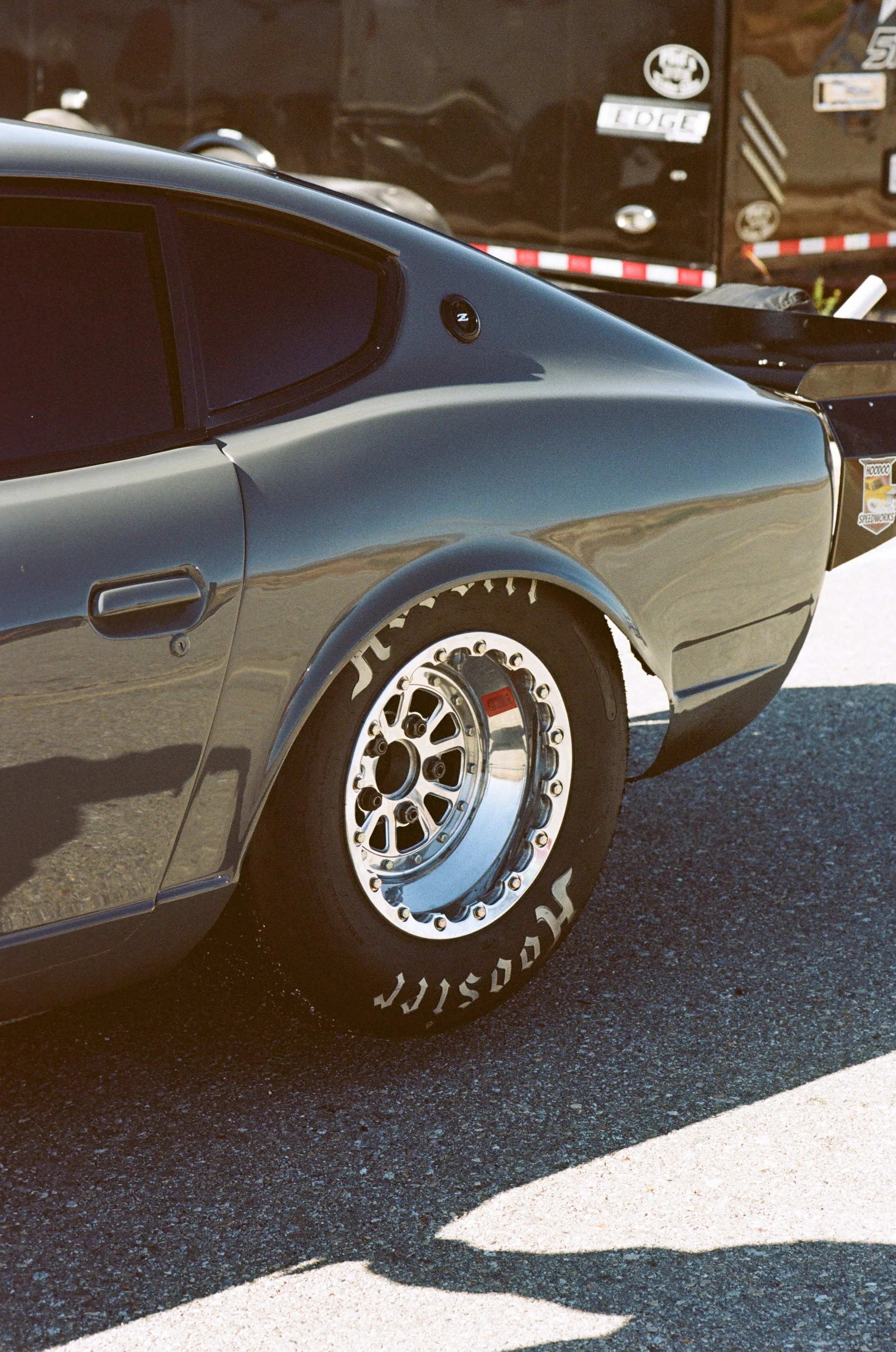 Close-up of a black vintage car's rear side, showing part of the wheel, tire with white lettering, and a reflective body in sunlight.