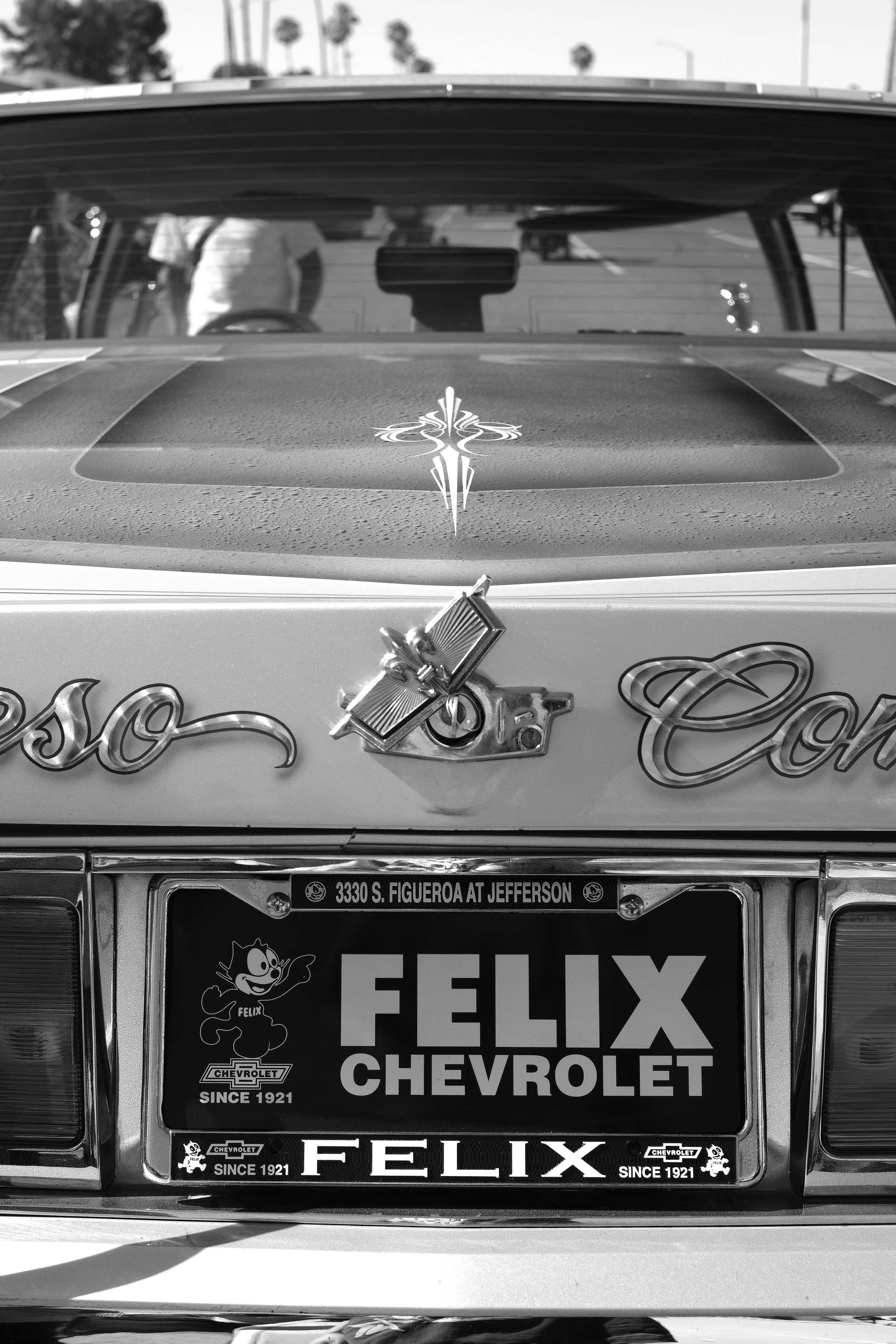 Rear view of a vintage Chevrolet car with dealership license plate frame reading 'Felix Chevrolet' at 3330 S. Figueroa at Jefferson, featuring a Felix cartoon mascot and Chevrolet logos, with a decorative emblem on the trunk.