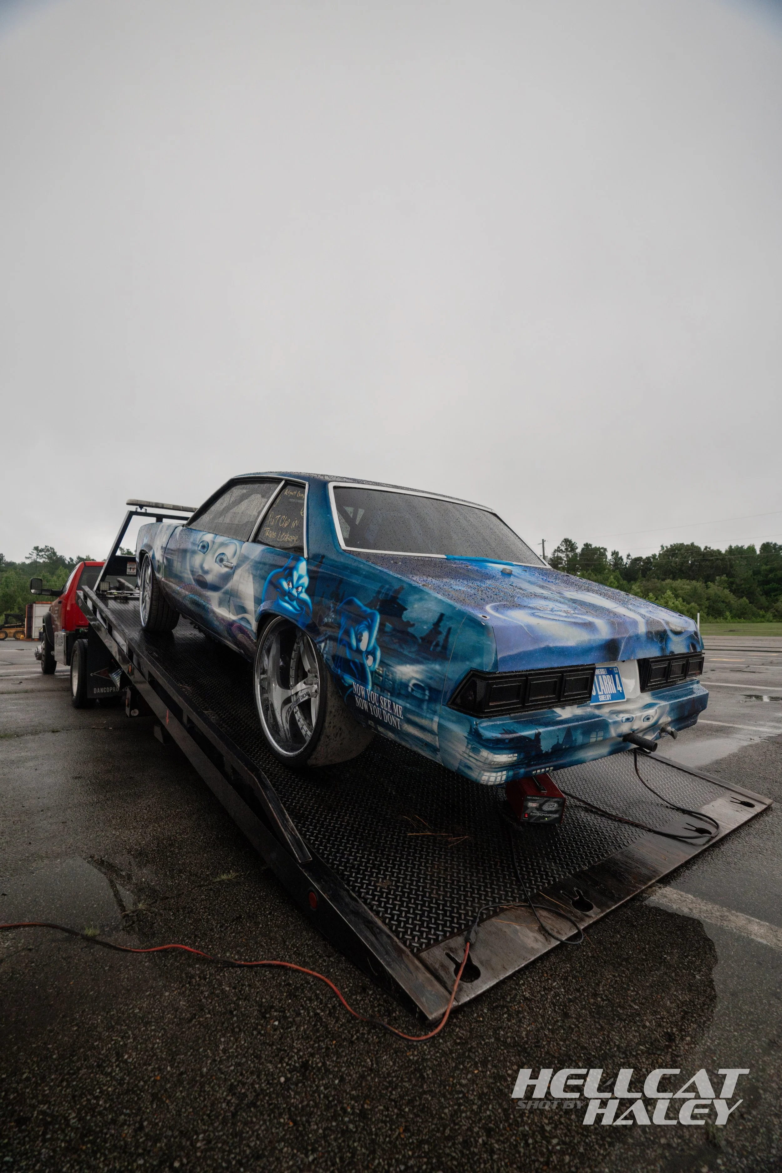 A blue race car with artwork on the sides, installed on a flatbed trailer, in a parking lot on a rainy day.