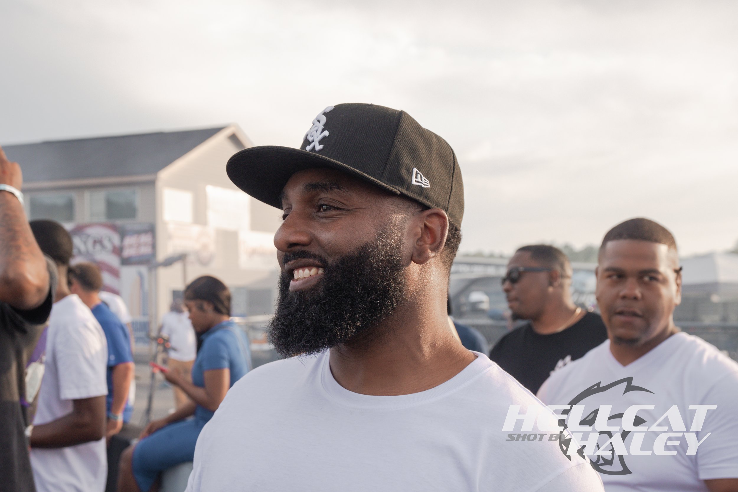 A man with a beard wearing a black sports cap and a white shirt at an outdoor gathering with people in the background.