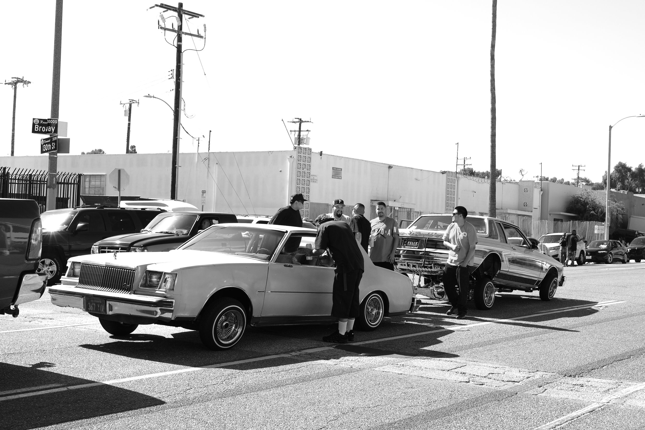 A group of people gathered around vintage cars in a parking lot with power lines and a building in the background.