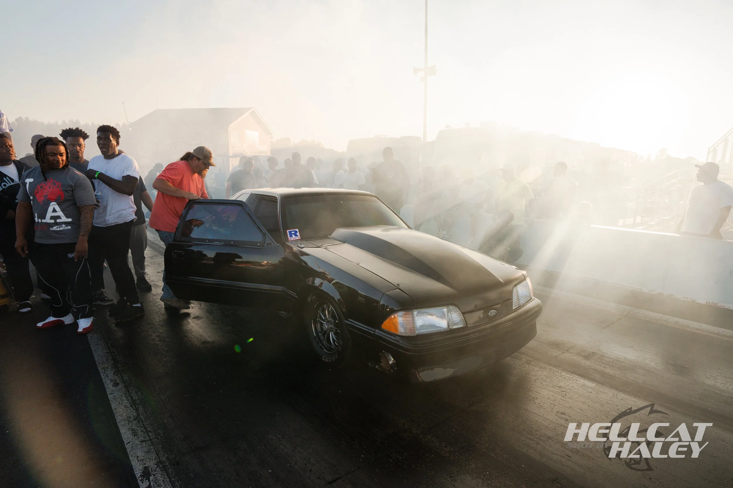 A group of people gathered around a black car on a smoky street at sunset.