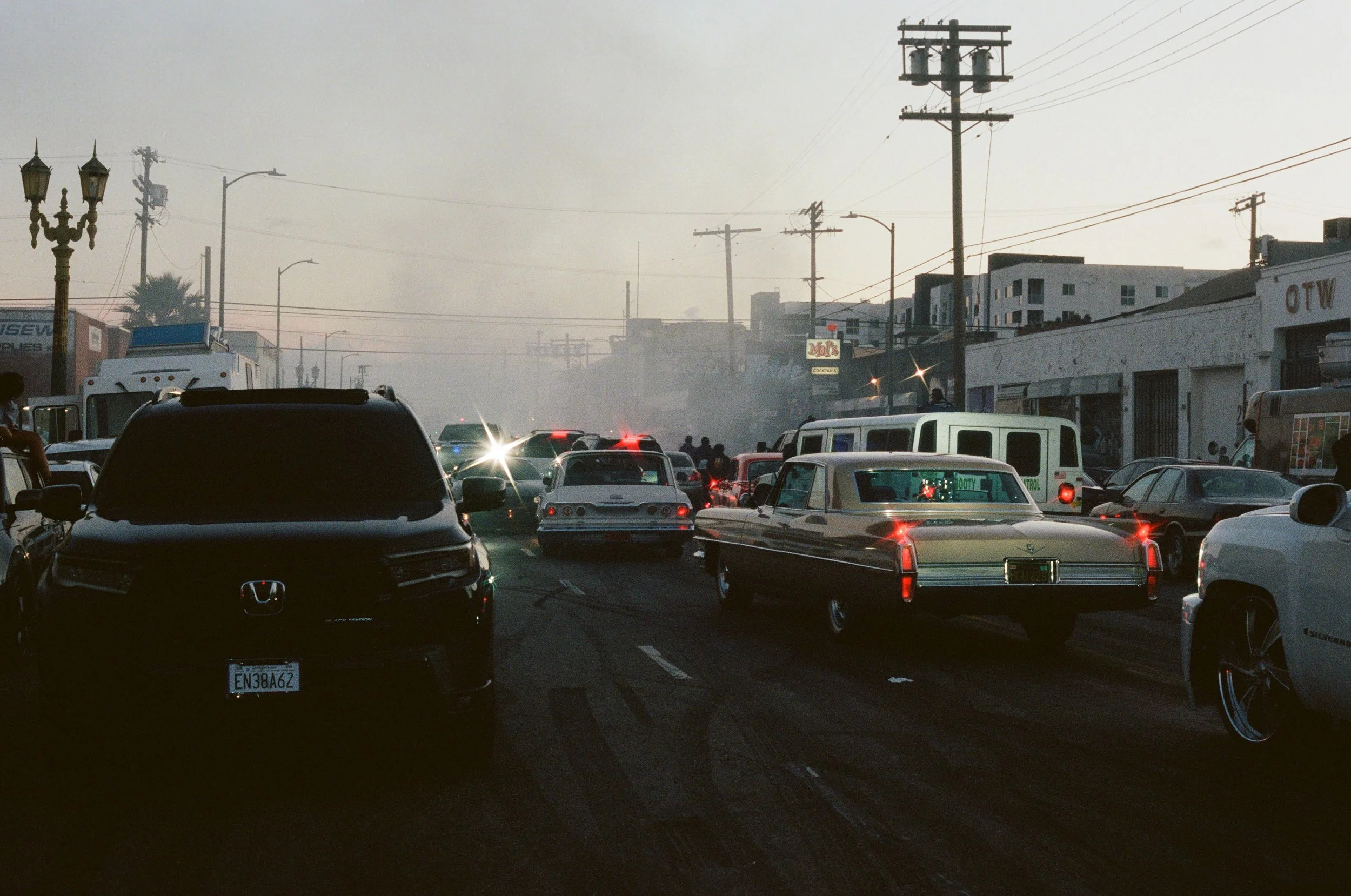 A busy parking lot with numerous cars and trucks, some with headlights on, under a hazy sky with power lines and buildings in the background.