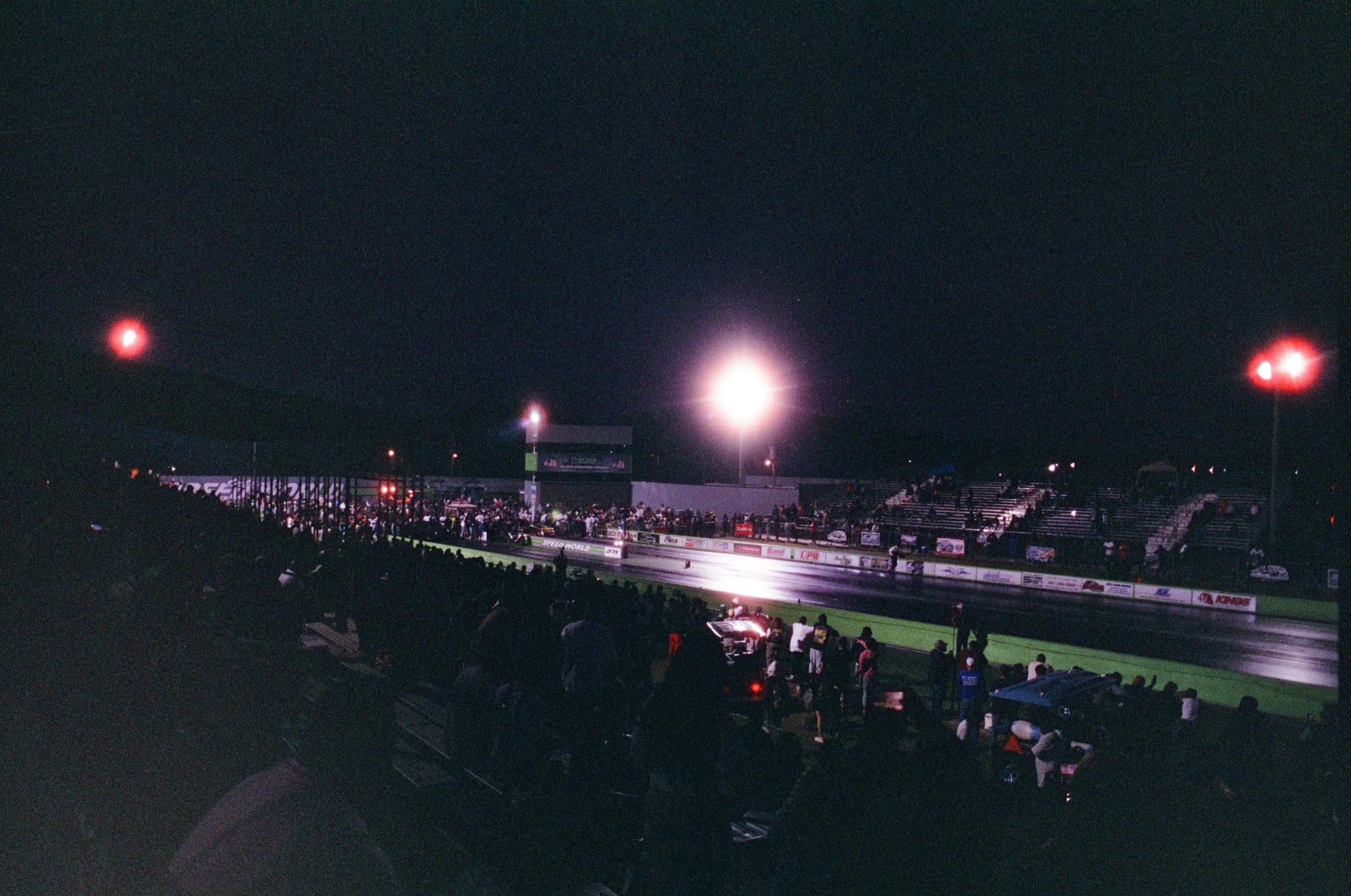Nighttime scene at a race track with spectators in the stands, bright stadium lights illuminating the track, and a few cars visible on the track.