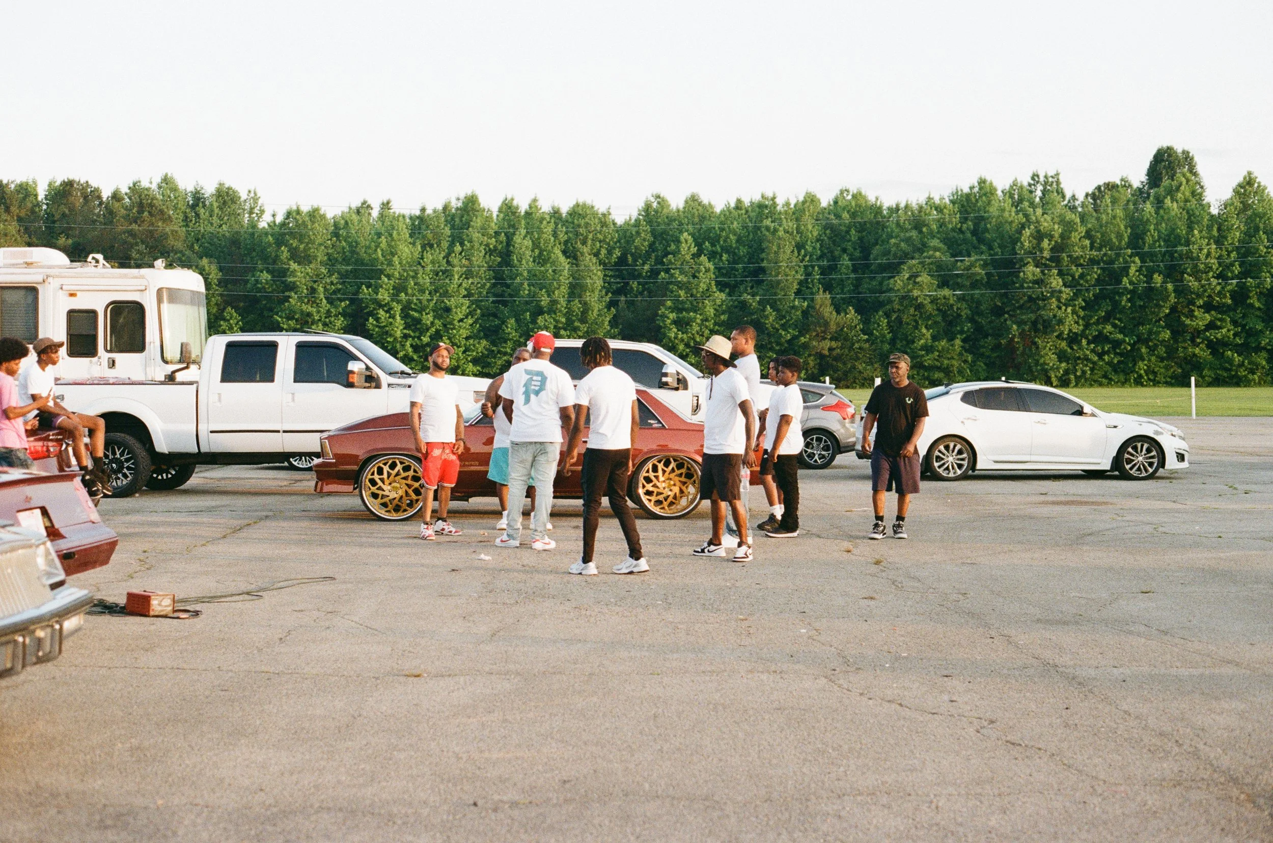 Group of people socializing outdoors in a parking lot with trees in the background, featuring various parked cars and a white camper trailer.