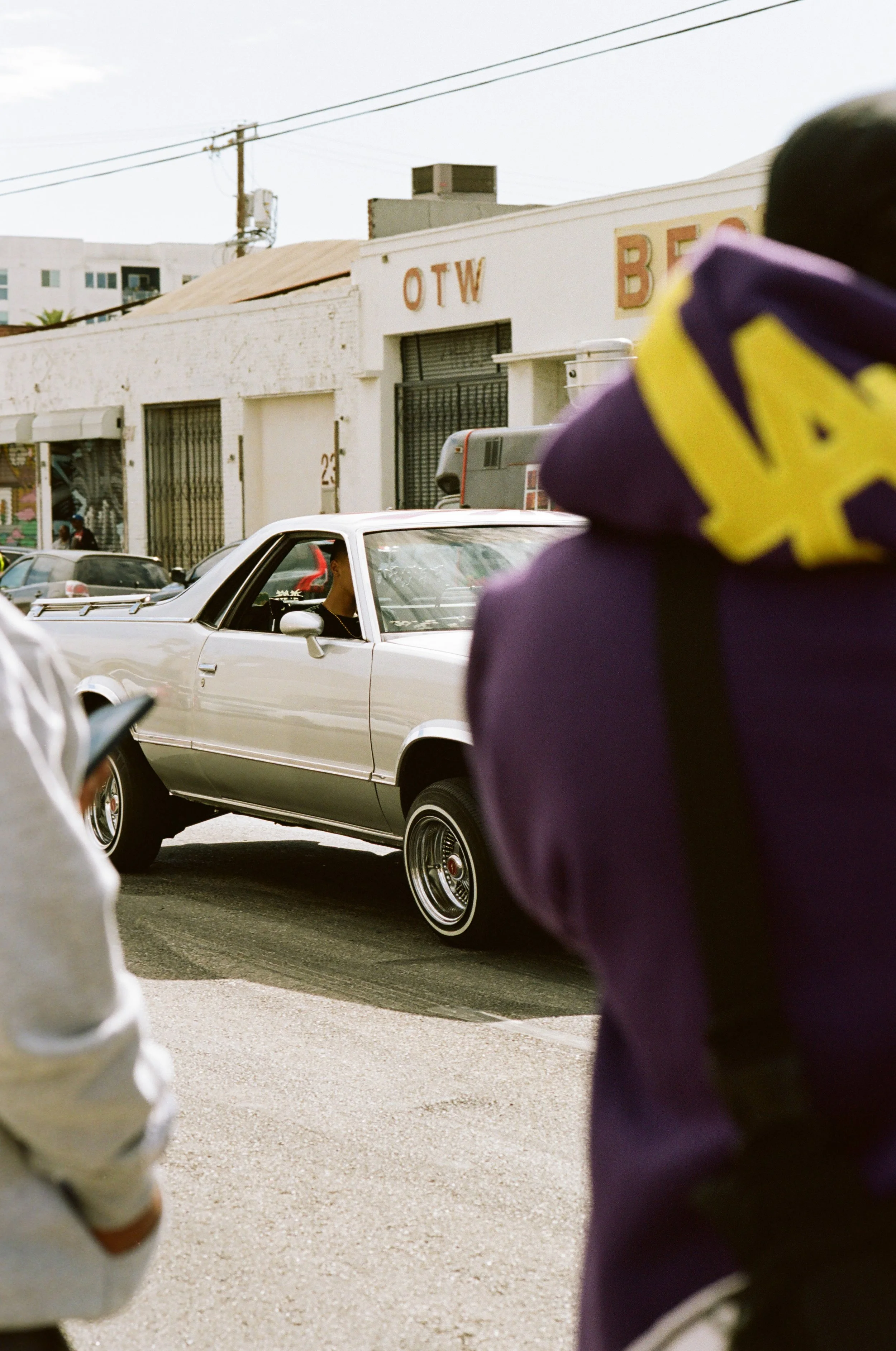 Street scene with a vintage white car in motion, two individuals visible in the foreground, and a building with signs reading 'OTW' and 'BE' in the background.