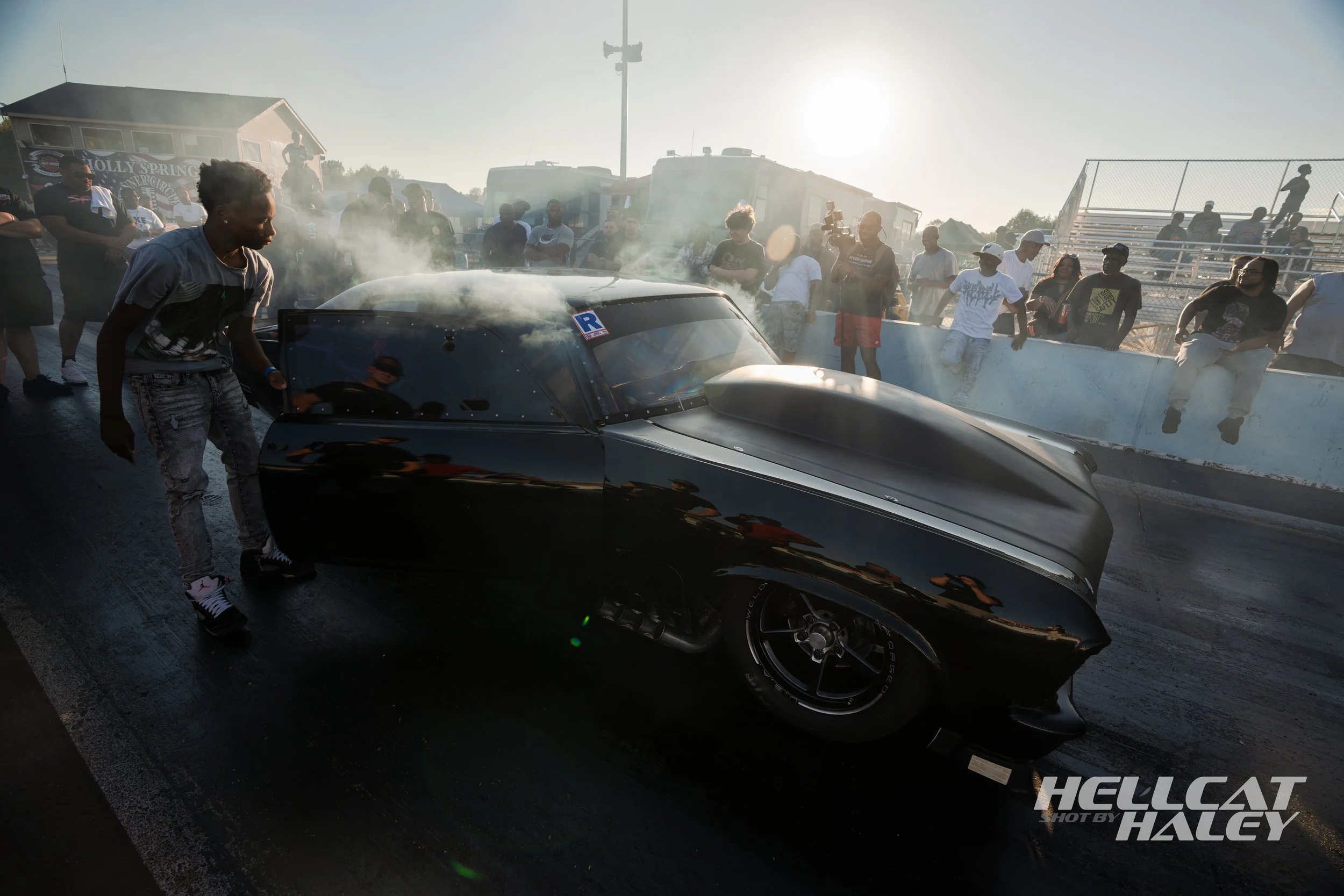 Black racing car at a drag strip with smoke coming from the rear tires as a crowd of spectators and crew members watch in the background during sunset.