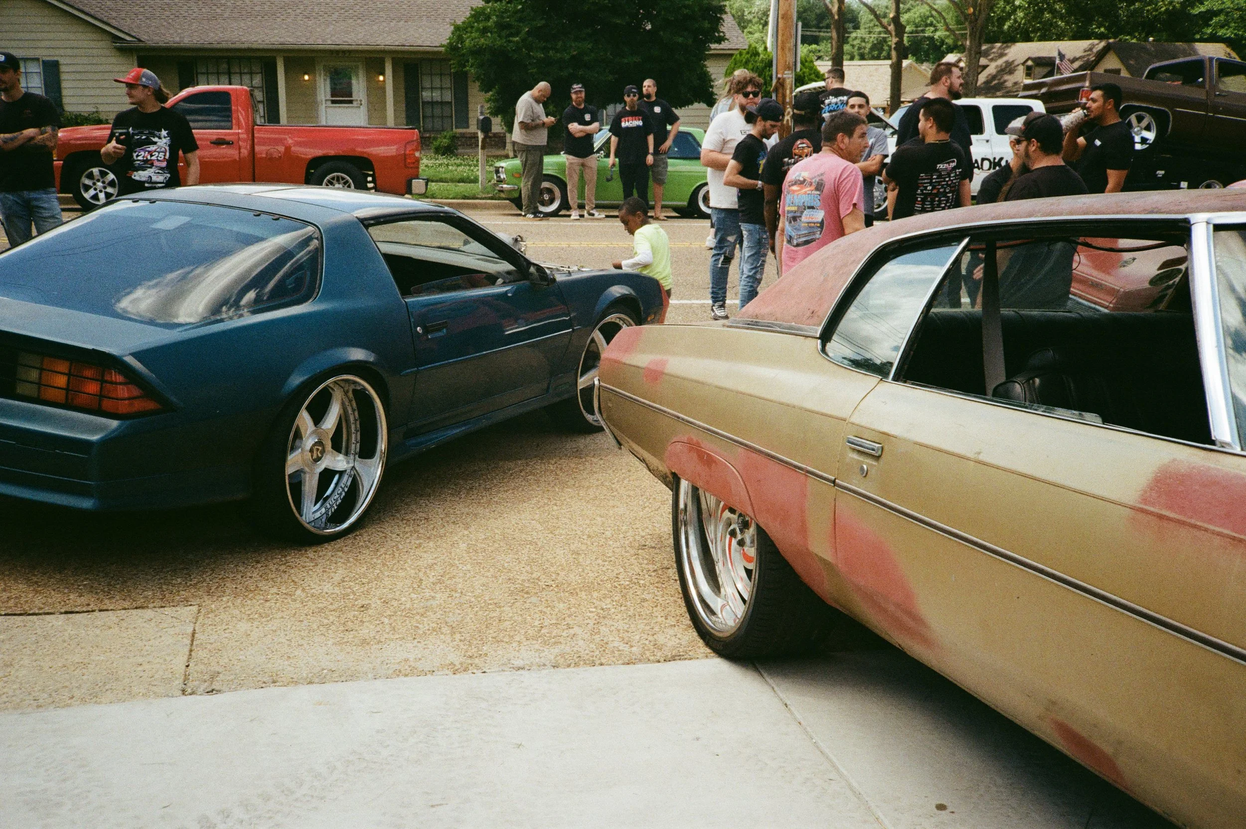 A group of people gathered outdoors during the daytime, surrounded by vintage and modern cars parked along a residential street with houses and trees in the background.