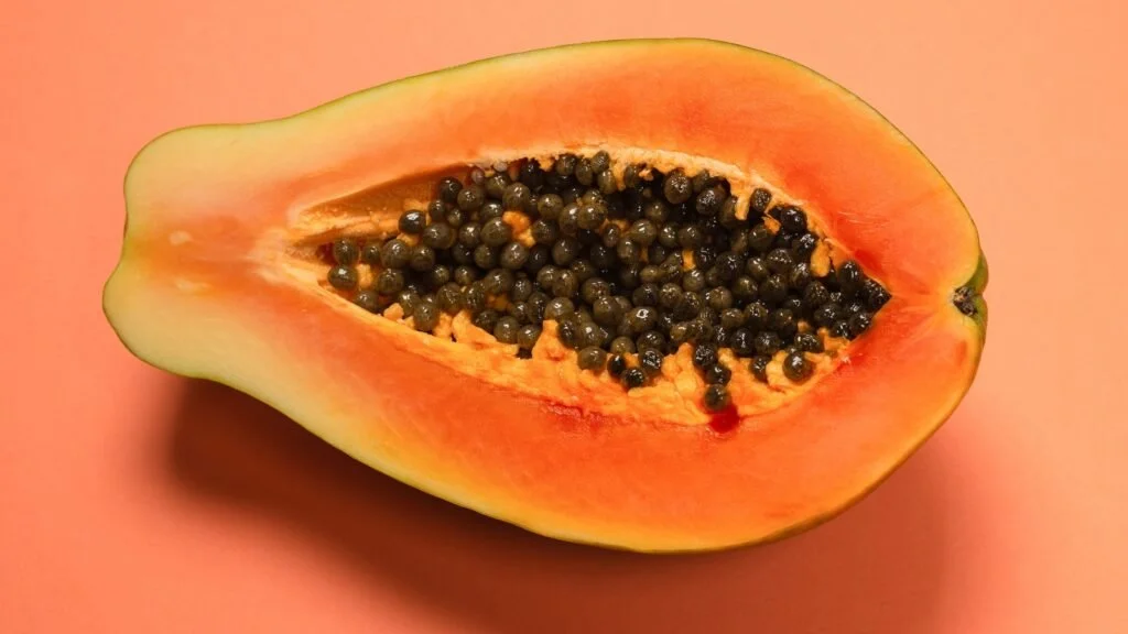 Half of a ripe papaya fruit with orange flesh and black seeds on a pink background.