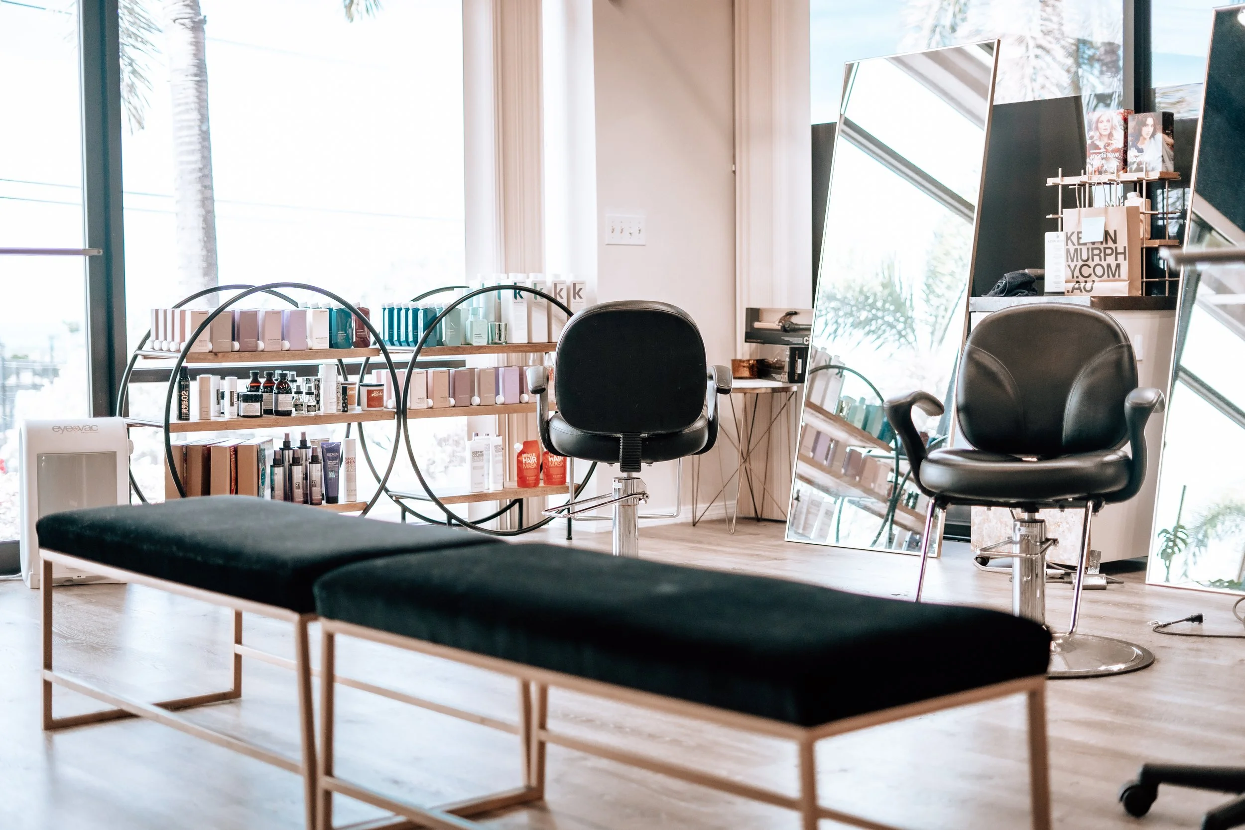 A modern salon interior with two black salon chairs, a large mirror, and shelves with hair and skincare products. There is a black upholstered bench in the foreground and a large window letting in natural light.