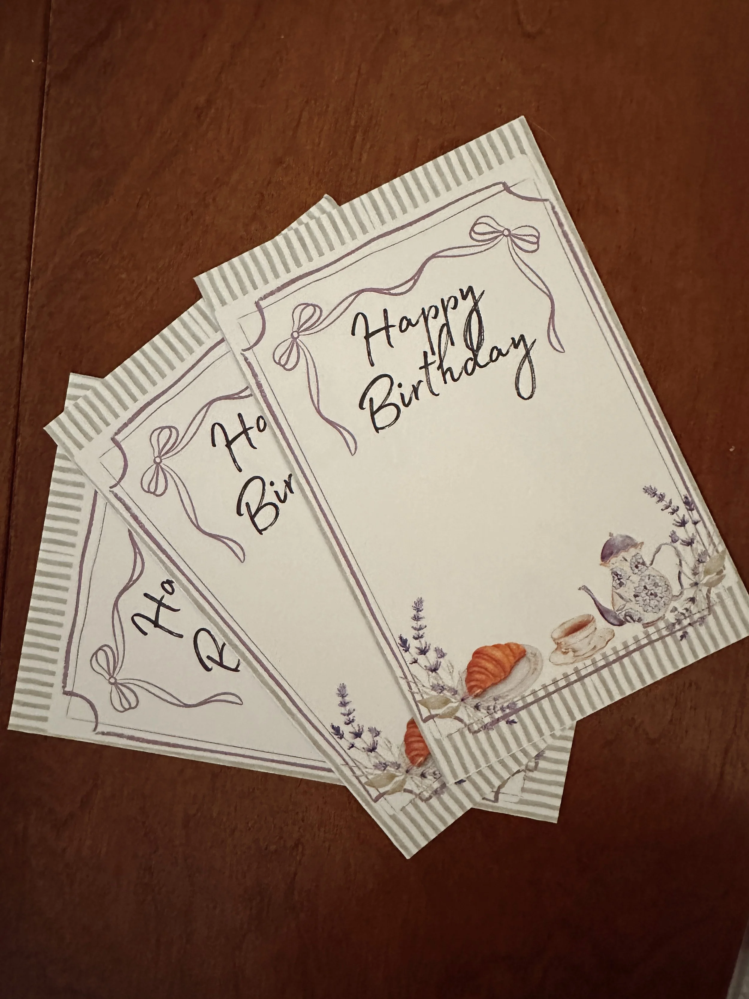 Three birthday cards with lavender and teapot illustrations on a wooden surface.