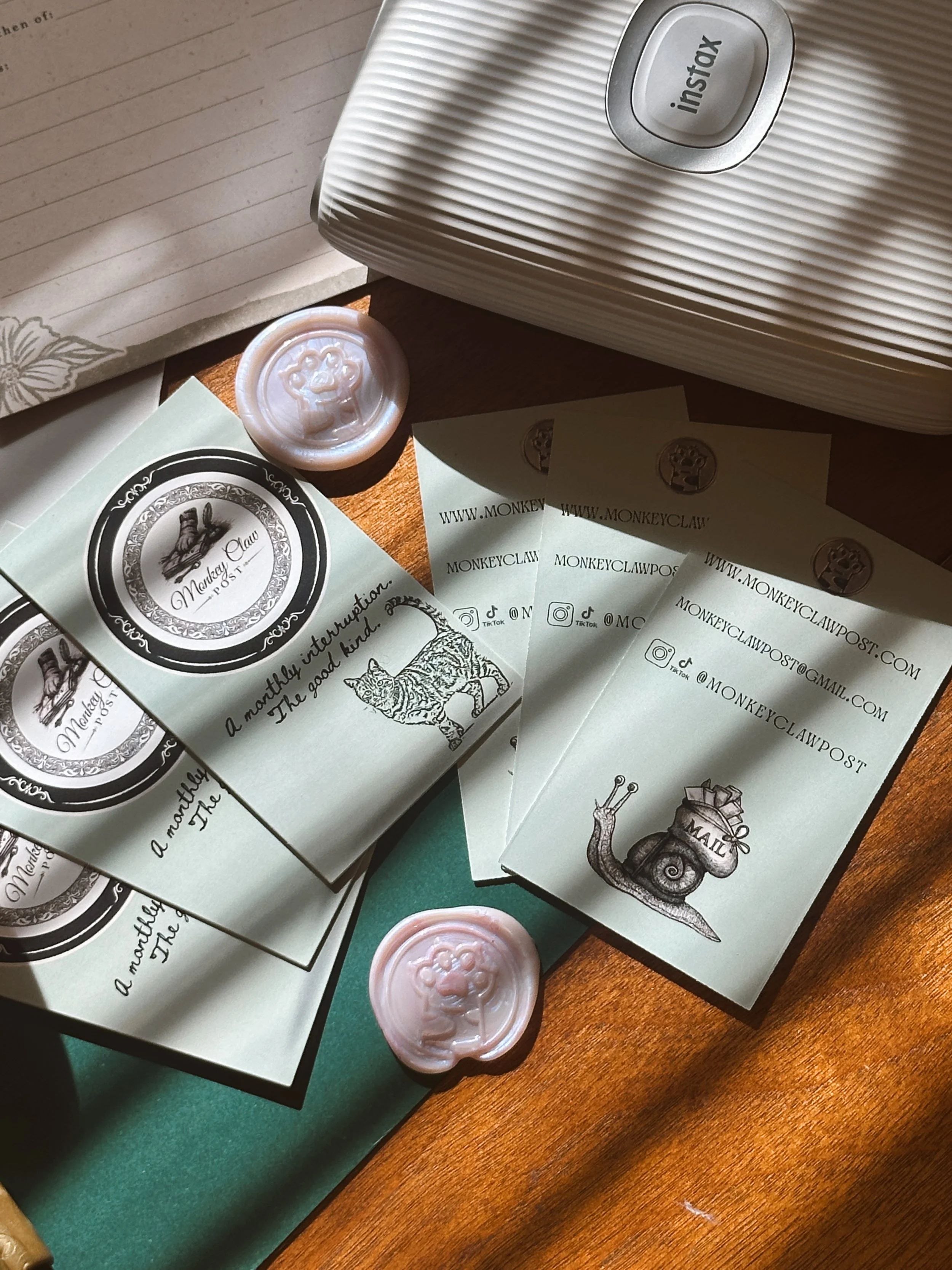 Business cards and wax seals from Monkey Claw. The cards display the logo, social media handles, and a snail illustration. The wax seals feature a playful lion face design. The scene includes a white Instax printer.