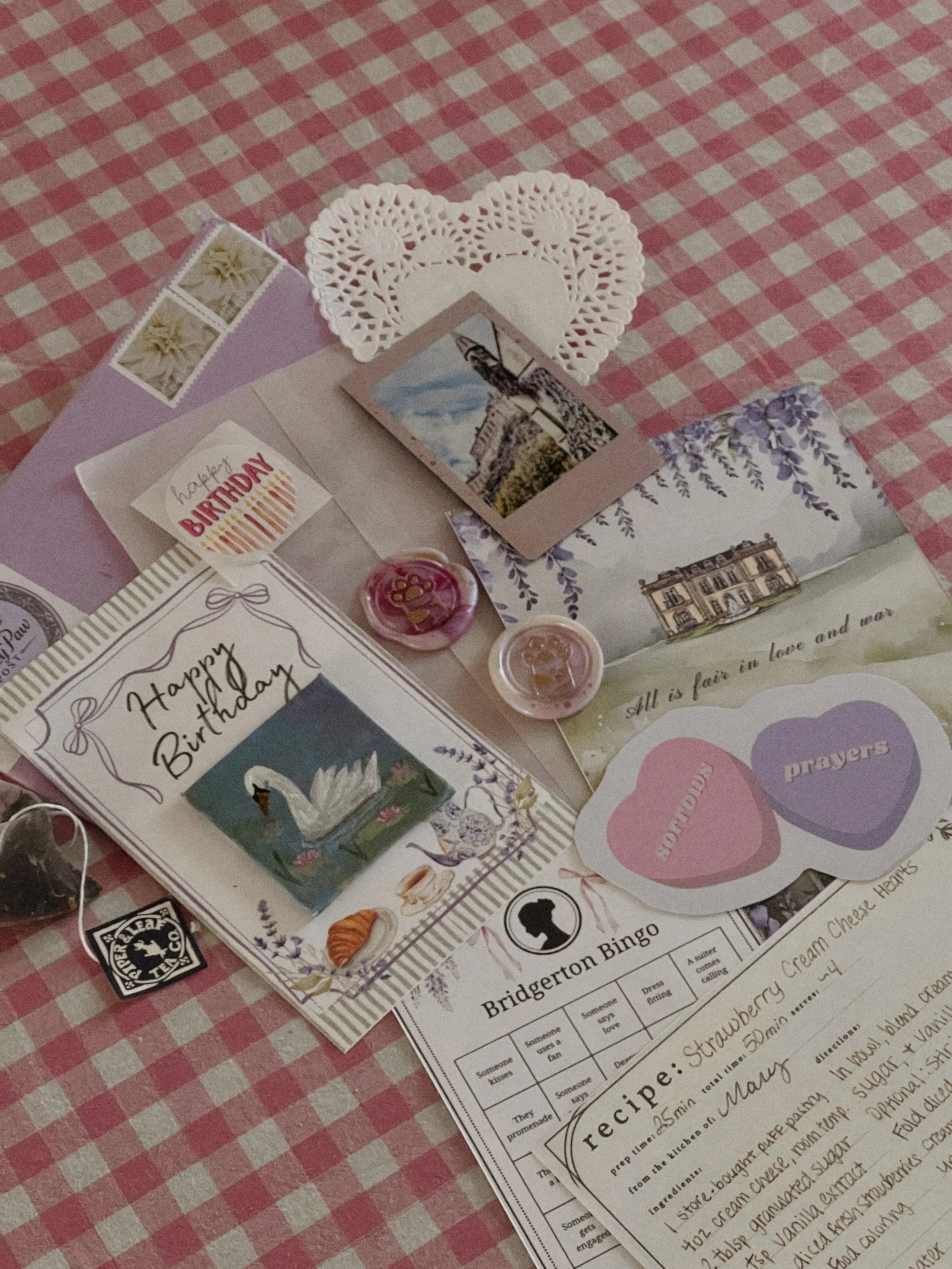 A birthday card and various decorative items on a pink and white checkered tablecloth. Included are a 'Happy Birthday' card with a swan illustration, small photo, two heart-shaped paper cutouts labeled 'Bountiful' and 'Prayers,' a bingo game sheet ti