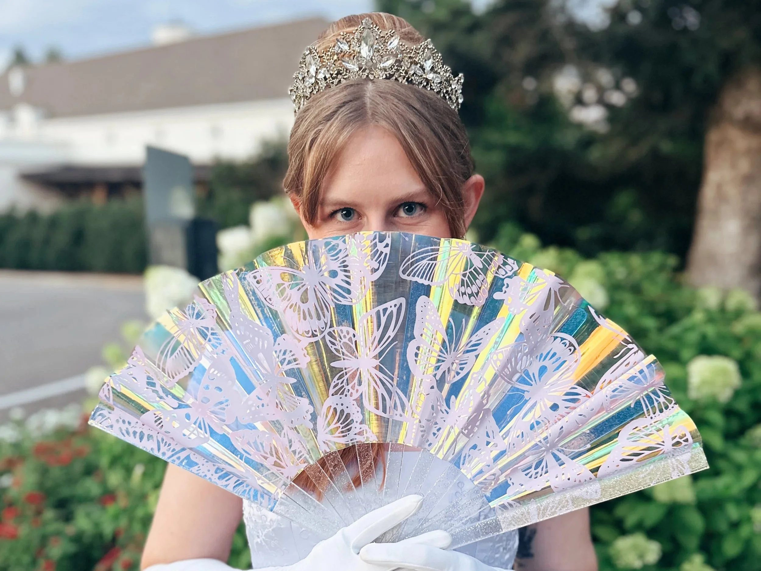 A woman wearing a tiara holds a decorative fan with butterfly cutouts in front of her face, eyes visible. She is outdoors with green foliage and trees in the background.