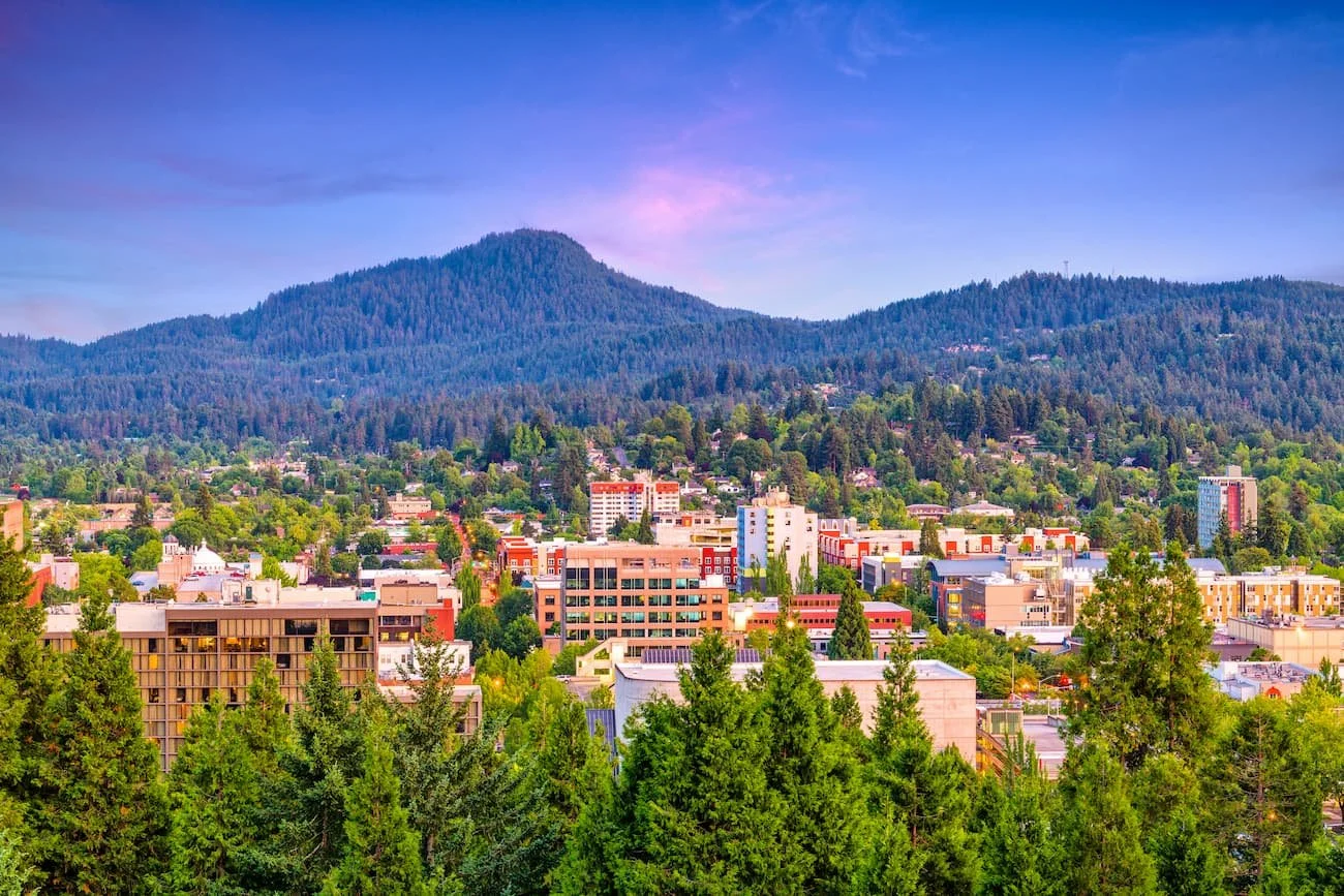 A cityscape with colorful buildings nestled among green trees and mountains in the background under a pink and purple sky.