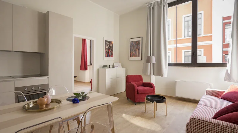 Living room with a red armchair, a couch with red pillows, a black side table, a white sideboard, and large windows with white curtains showing an orange building outside.