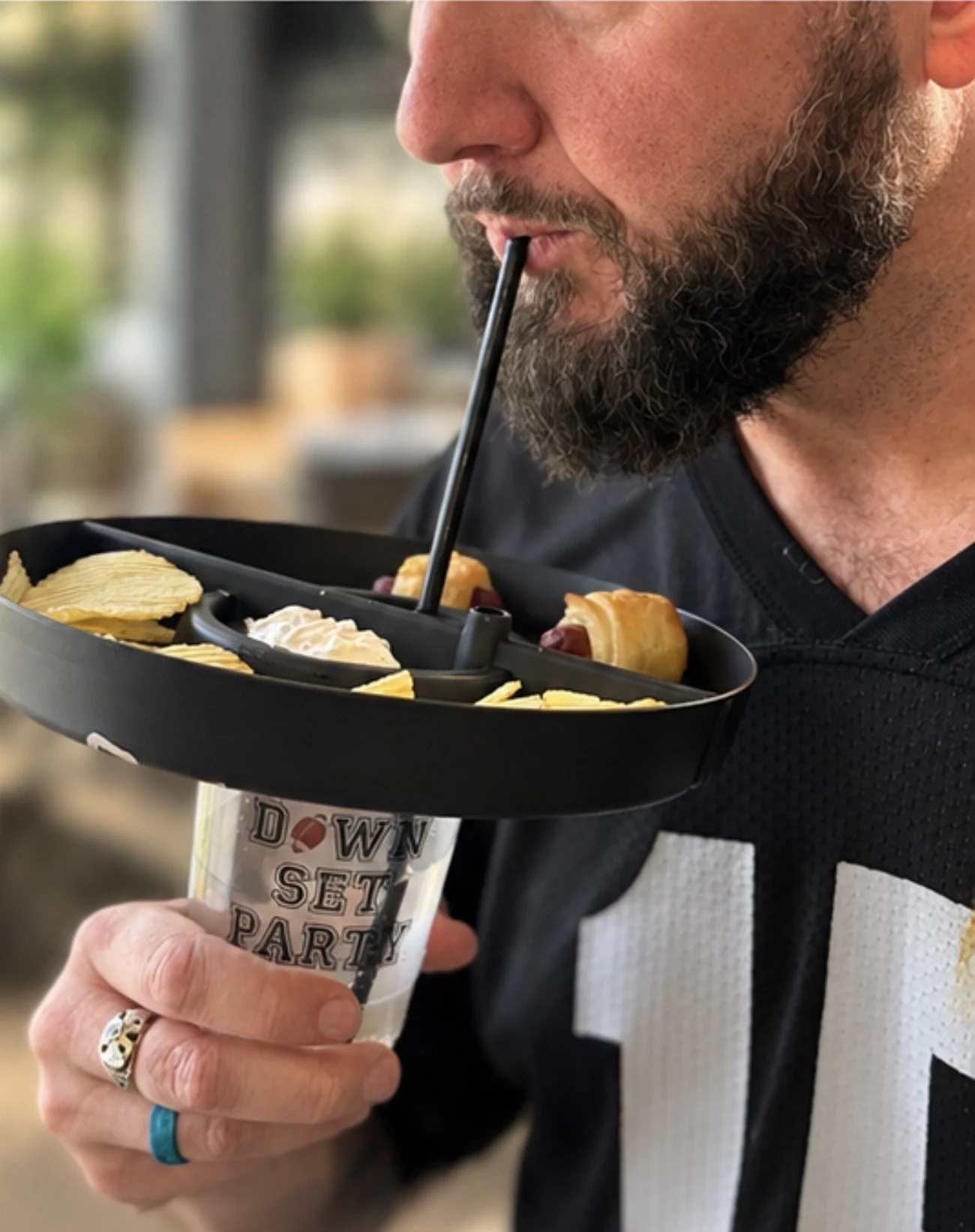 A man with a beard drinking from a straw inserted into a tray of potato chips and hot dog sandwiches, held over a cup with the text 'Downset Party'.