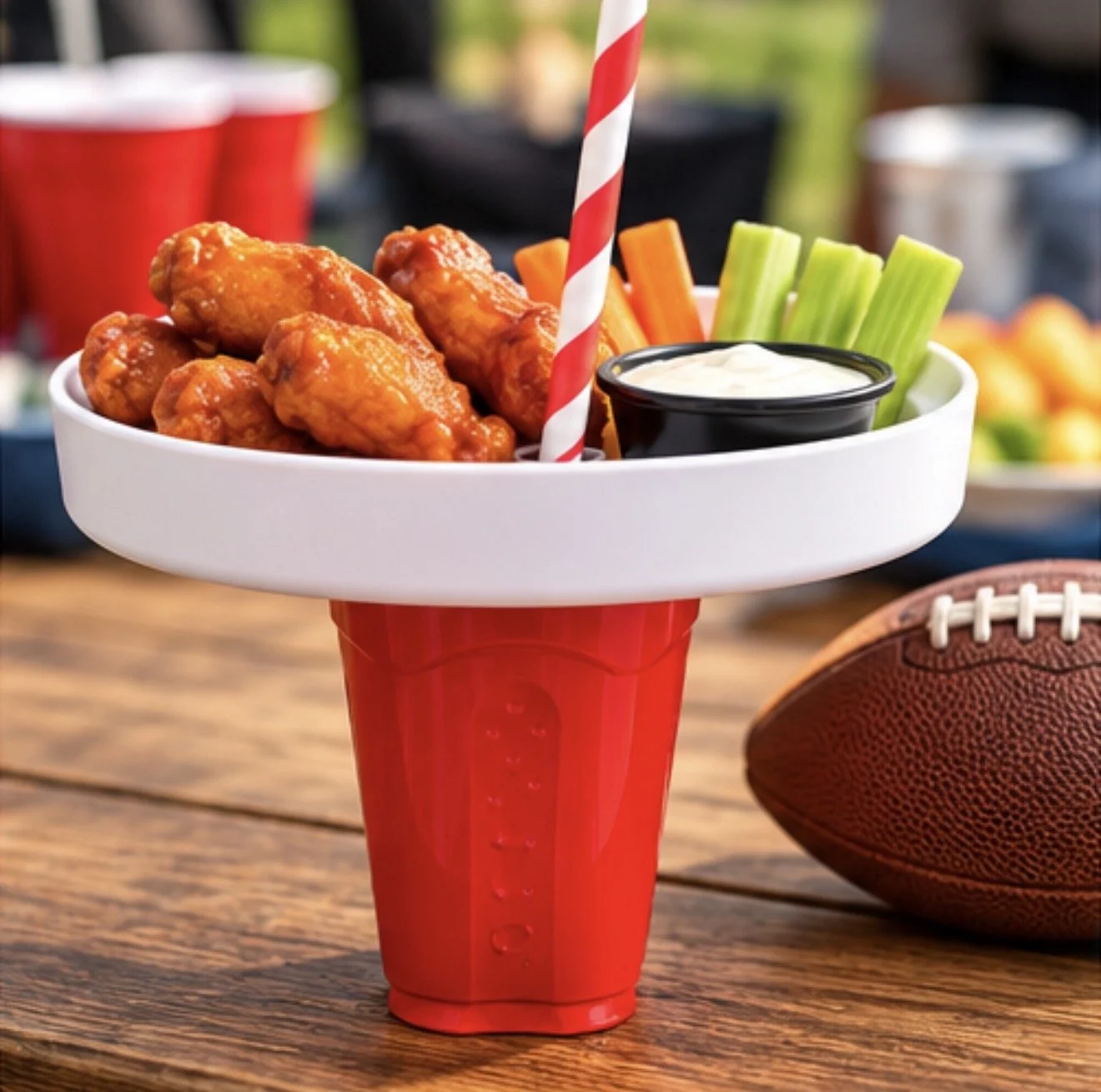 A football-shaped snack tray on a red cup with buffalo chicken wings, celery sticks, carrots, and a side of ranch dressing. A striped straw is in the cup. In the background, there are red cups, bowls of snacks, and a football on a wooden table.