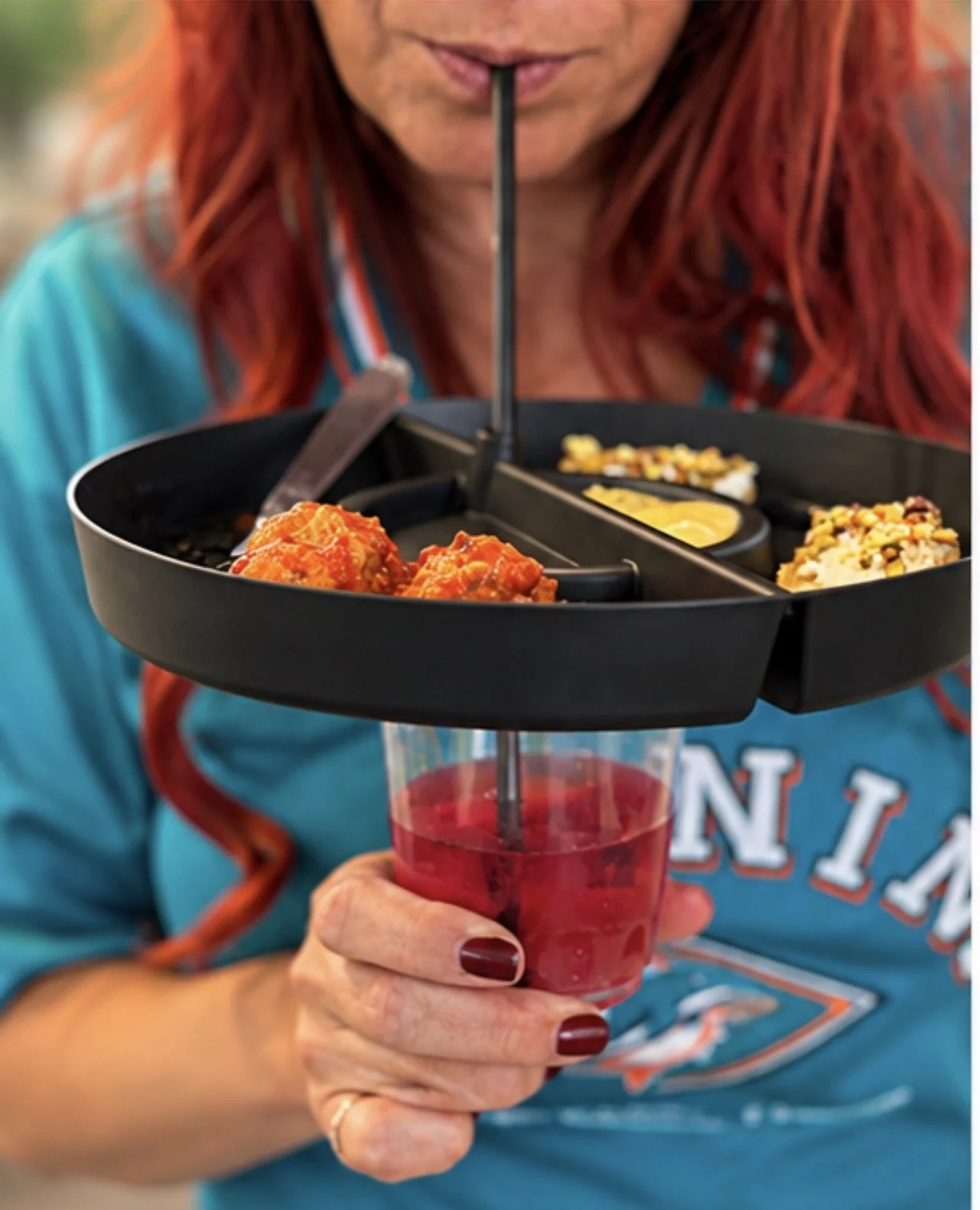 A woman with red hair holding a red drink with a black tiered snack holder with various snacks and dips above the glass.