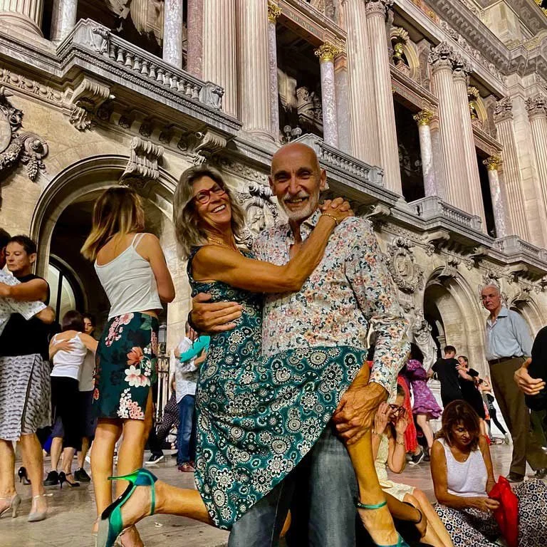 A woman happily dancing together at a historic building with marble columns and intricate architecture. The woman has her arms around the man's neck, and they are smiling. Several other people are in the background, some dancing and some sitting.