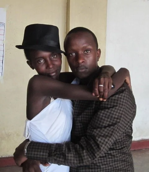 Two young people embrace each other indoors, one wearing a black hat and white sleeveless shirt, the other wearing a plaid jacket. The background has a cream-colored wall and a partially visible whiteboard.