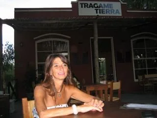 Woman with brown hair sitting at a table outside of a brick building with a sign that says 'TRAGAME TIERRA'
