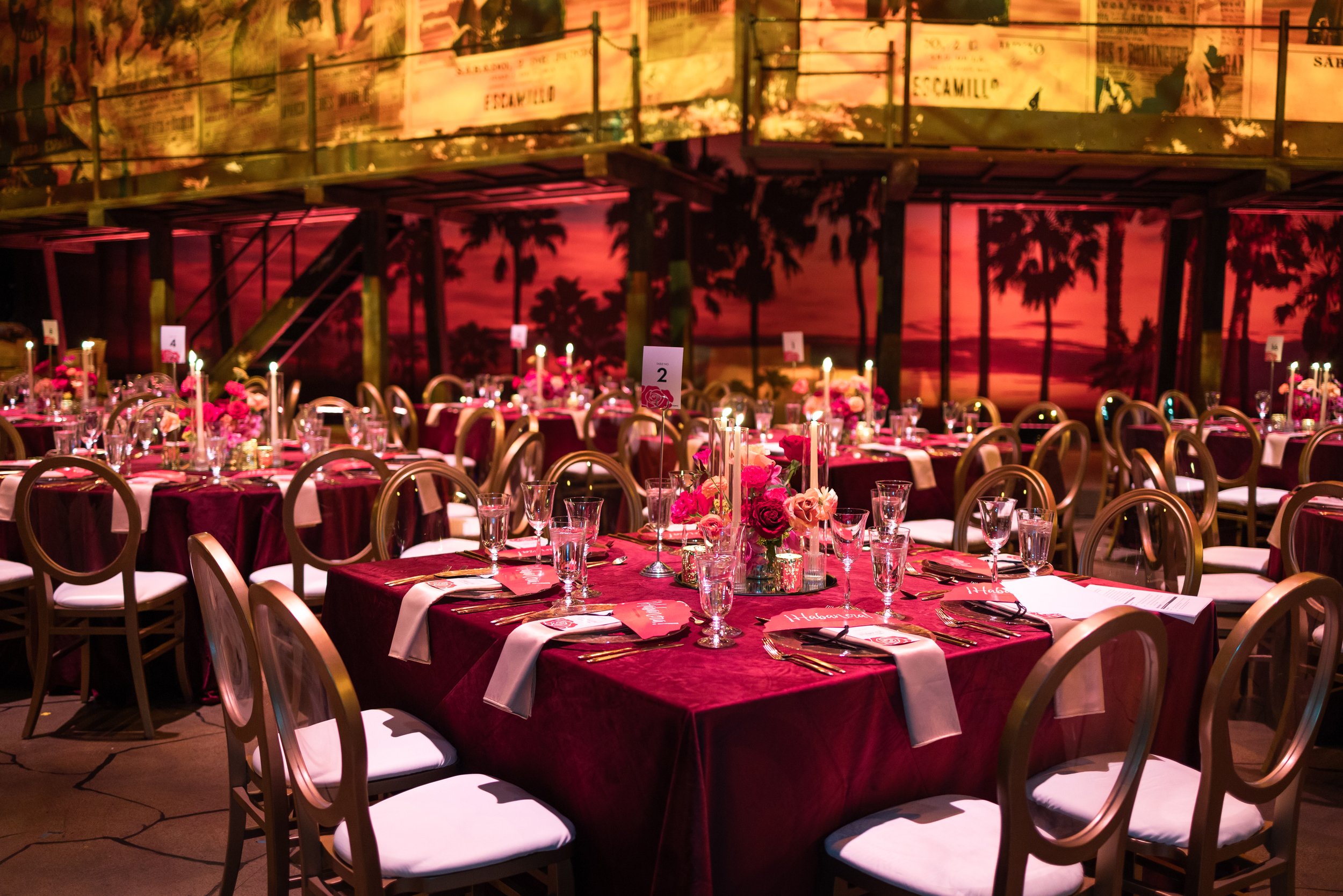 Velluto Events Co. Corporate Gala. A banquet hall decorated for an event with round tables covered in dark red velvet tablecloths, each set with glassware, gold utensils, and pink napkins. The tables have floral centerpieces with pink and red flowers