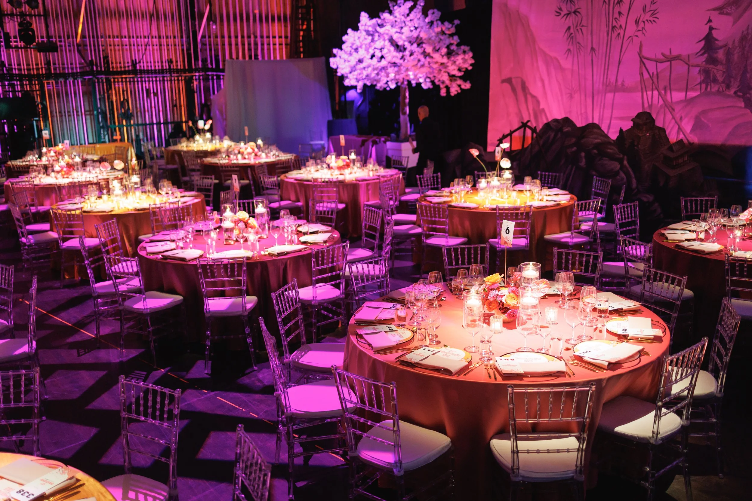 Velluto Events Co.  Corporate Event Elegant banquet hall with round tables covered in burgundy tablecloths, decorated with candles and floral centerpieces, and transparent chairs. Pink and purple lighting create a festive atmosphere, with a decorativ