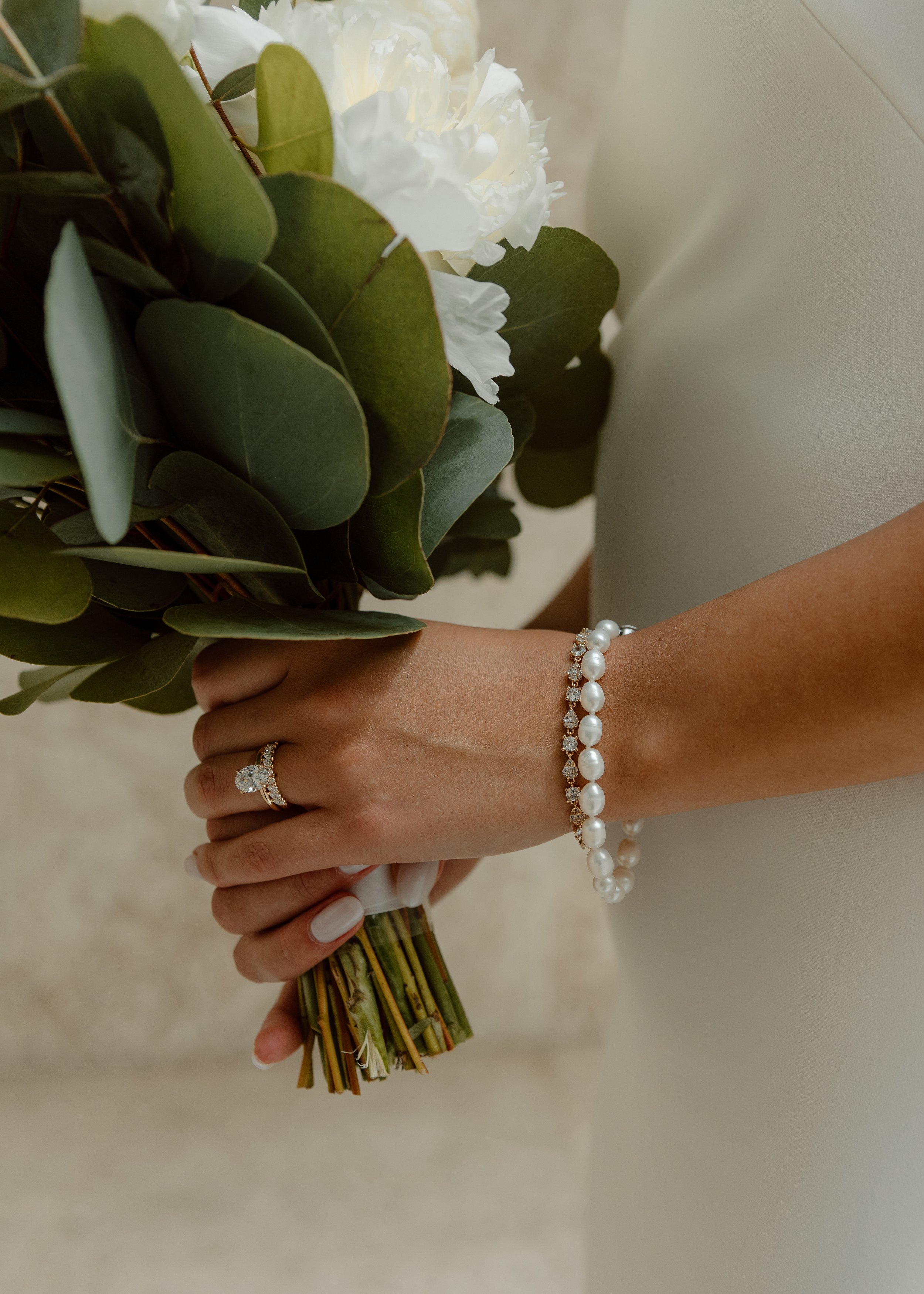 Velluto Events Co. Close-up of a woman's hand holding a bouquet of white flowers and green leaves, with a pearl bracelet and a diamond engagement ring visible.