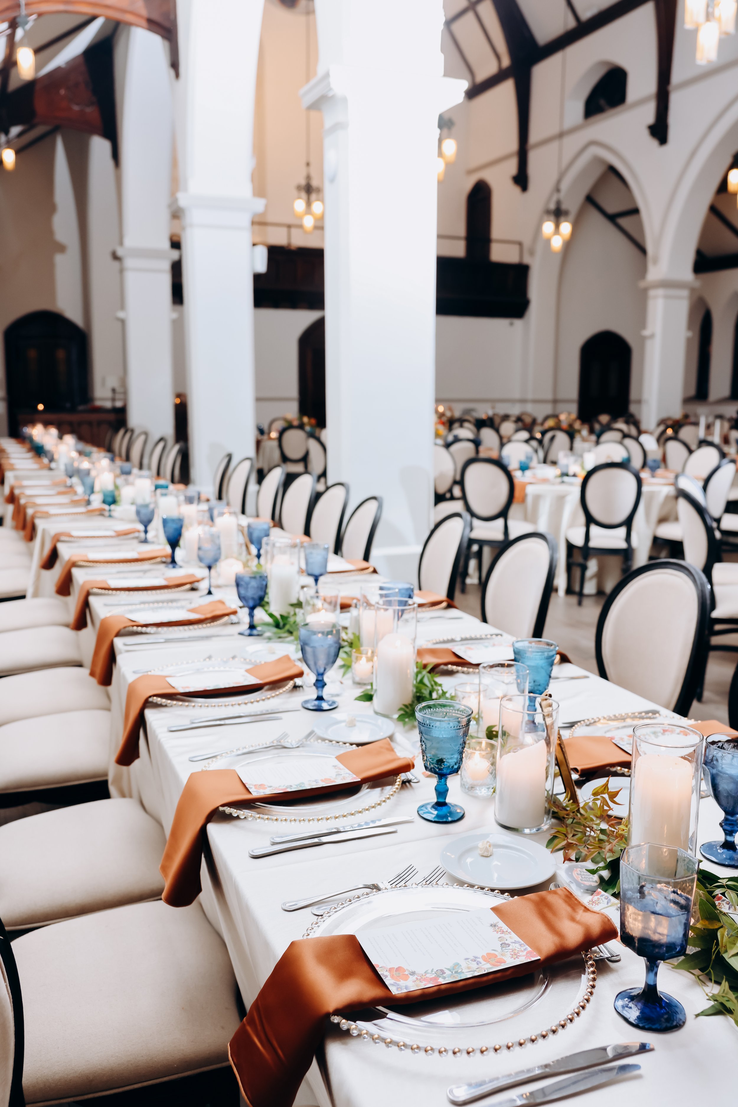 Velluto Events Co.  Wedding - Long banquet table set for a formal event in a grand hall with high, arched ceilings, white pillars, and hanging lights. The table is decorated with candles, greenery, blue goblets, and place settings with napkins and me