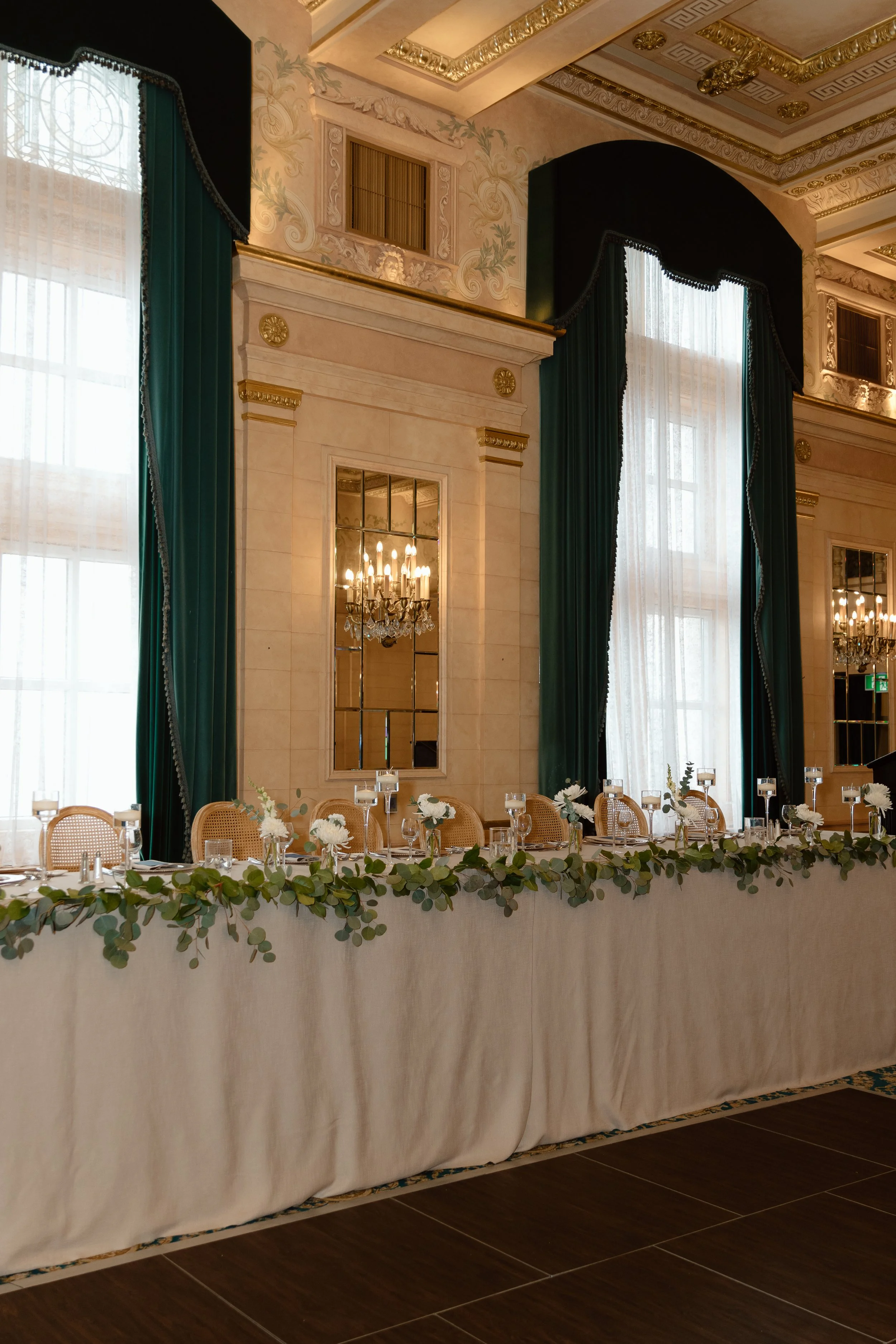 Velluto Events Co. Wedding elegant banquet table with white tablecloth, floral centerpieces, glassware, and candles in a grand decorated room with tall windows, green curtains, chandeliers, and ornate ceiling details.