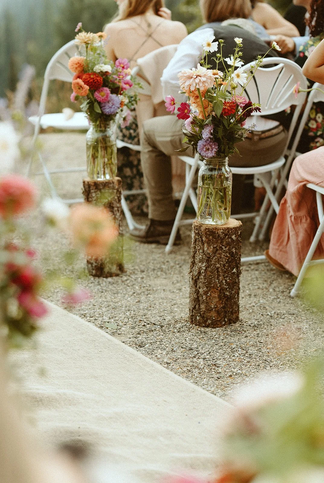 Velluto Events Co. Wedding aisle, vases with colorful flowers resting on tree stumps, set up for an outdoor event with seated guests in the background.