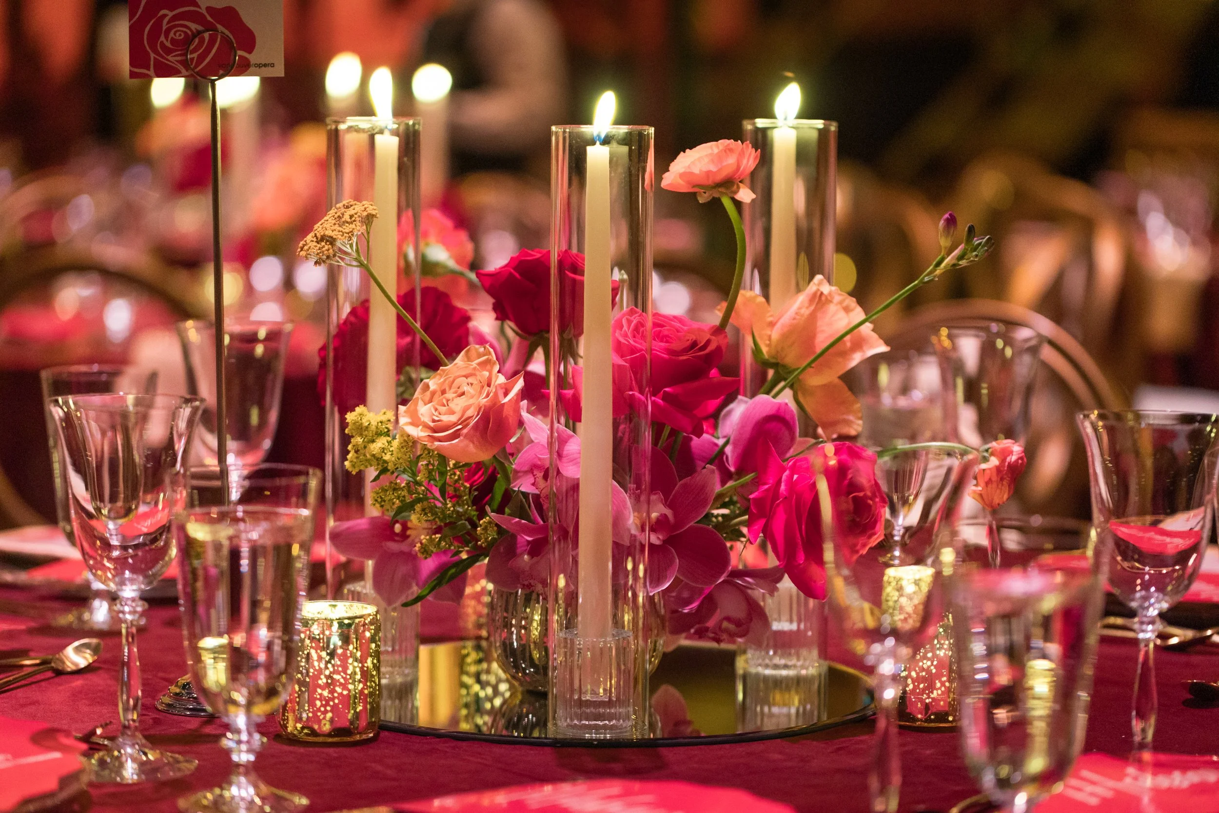 Velluto Events Co. Corporate Gala - A table setting with pink and red roses, orchids, and other flowers in tall glass vases, surrounded by glassware, candles, and a mirror base, with a blurred background and warm lighting.