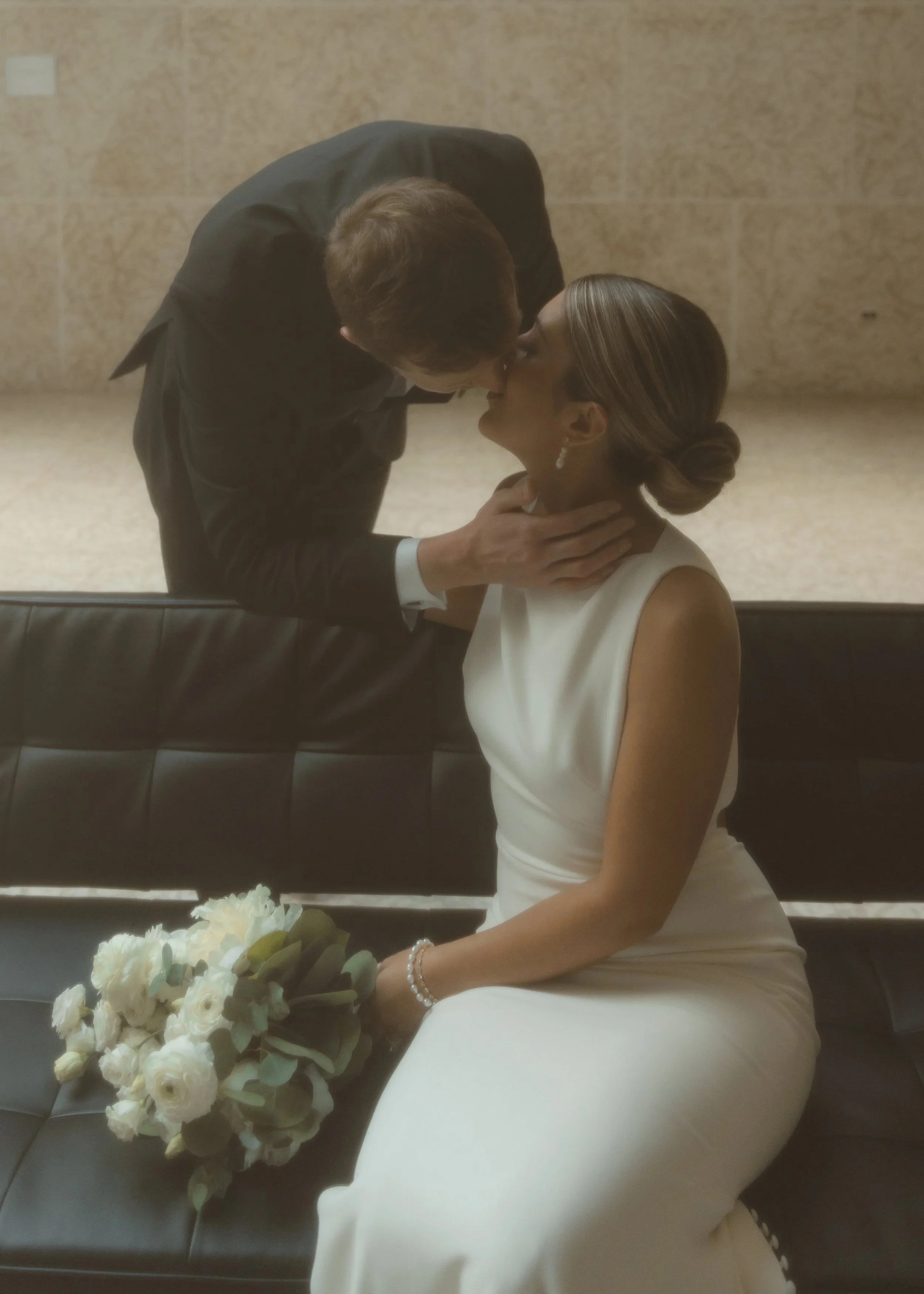 Velluto Events Co. A bride in a white wedding dress seated on a black bench, holding a bouquet, with a groom leaning in to kiss her.