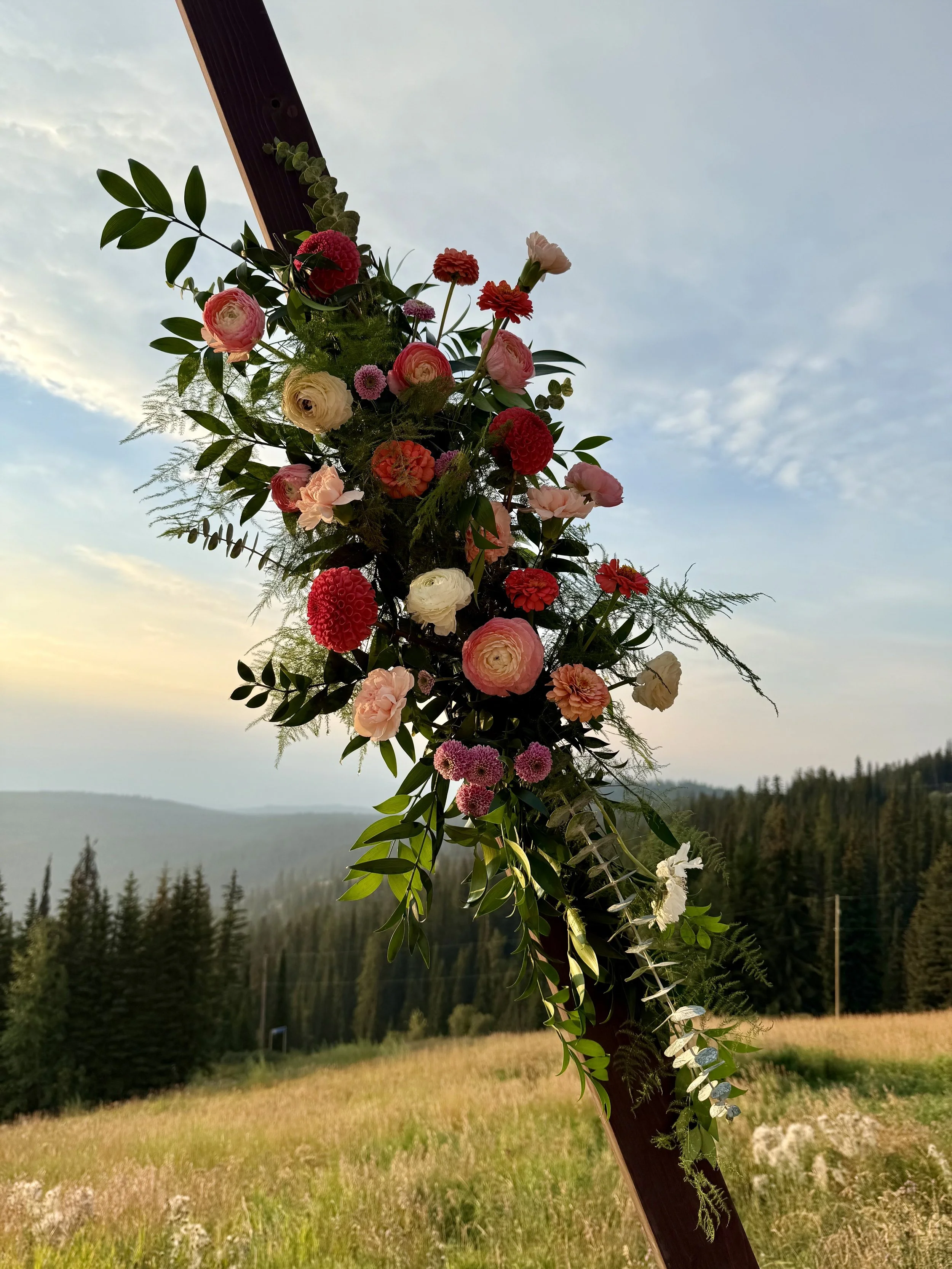 Velluto Events Co. Wedding arch decorated with various pink, red, and white flowers, set against a rural landscape with trees and hills at sunset.