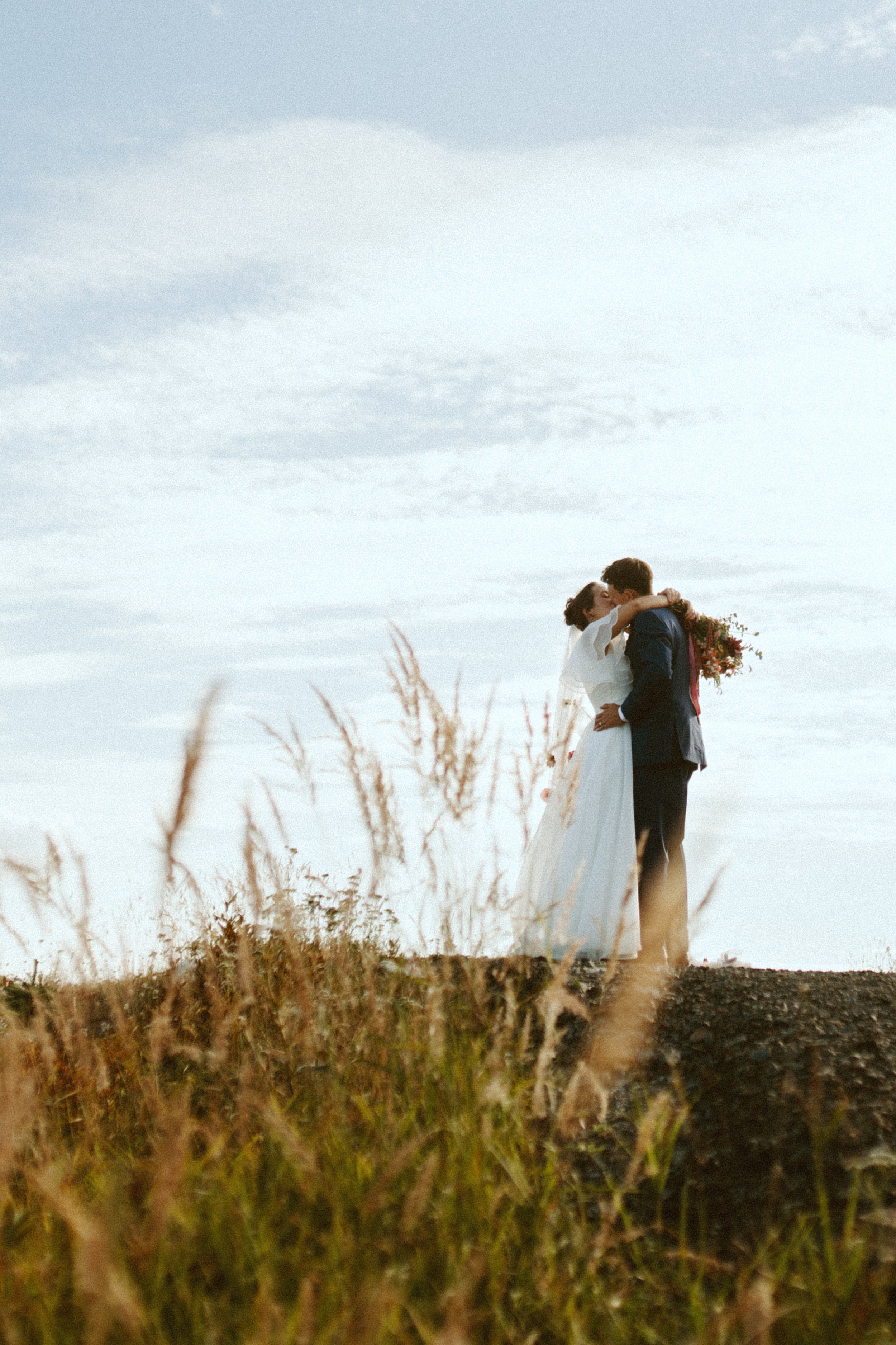 Velluto Events Co. A bride and groom sharing a kiss outdoors on an elevated ground with tall grass in the foreground, cloudy sky background.
