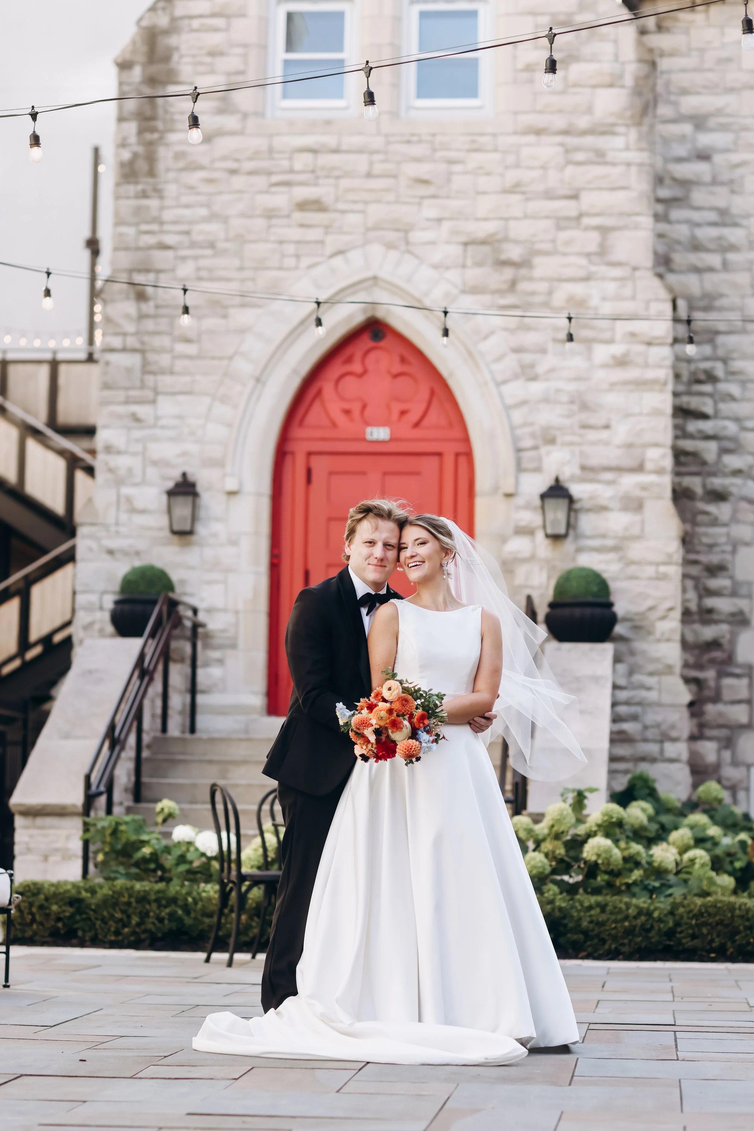 Velluto Events Co. Wedding A bride and groom in wedding attire smiling and embracing outdoors in front of a stone building with a red door, decorated with string lights and greenery.