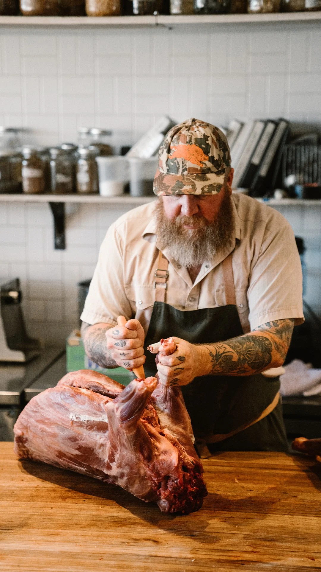 A man with tattoos and a beard, wearing a camouflage hat and an apron, butchering a large piece of meat on a wooden cutting board in a kitchen or butcher shop.