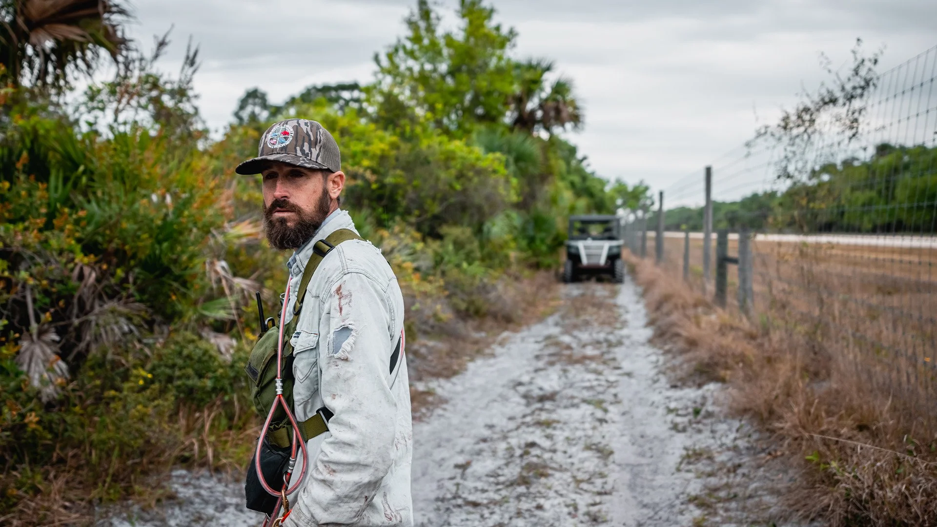 A man with a beard, wearing a baseball cap, a light-colored jacket, and a harness, standing on a dirt path bordered by a fence on one side and dense bushes on the other, with an off-road utility vehicle in the background.