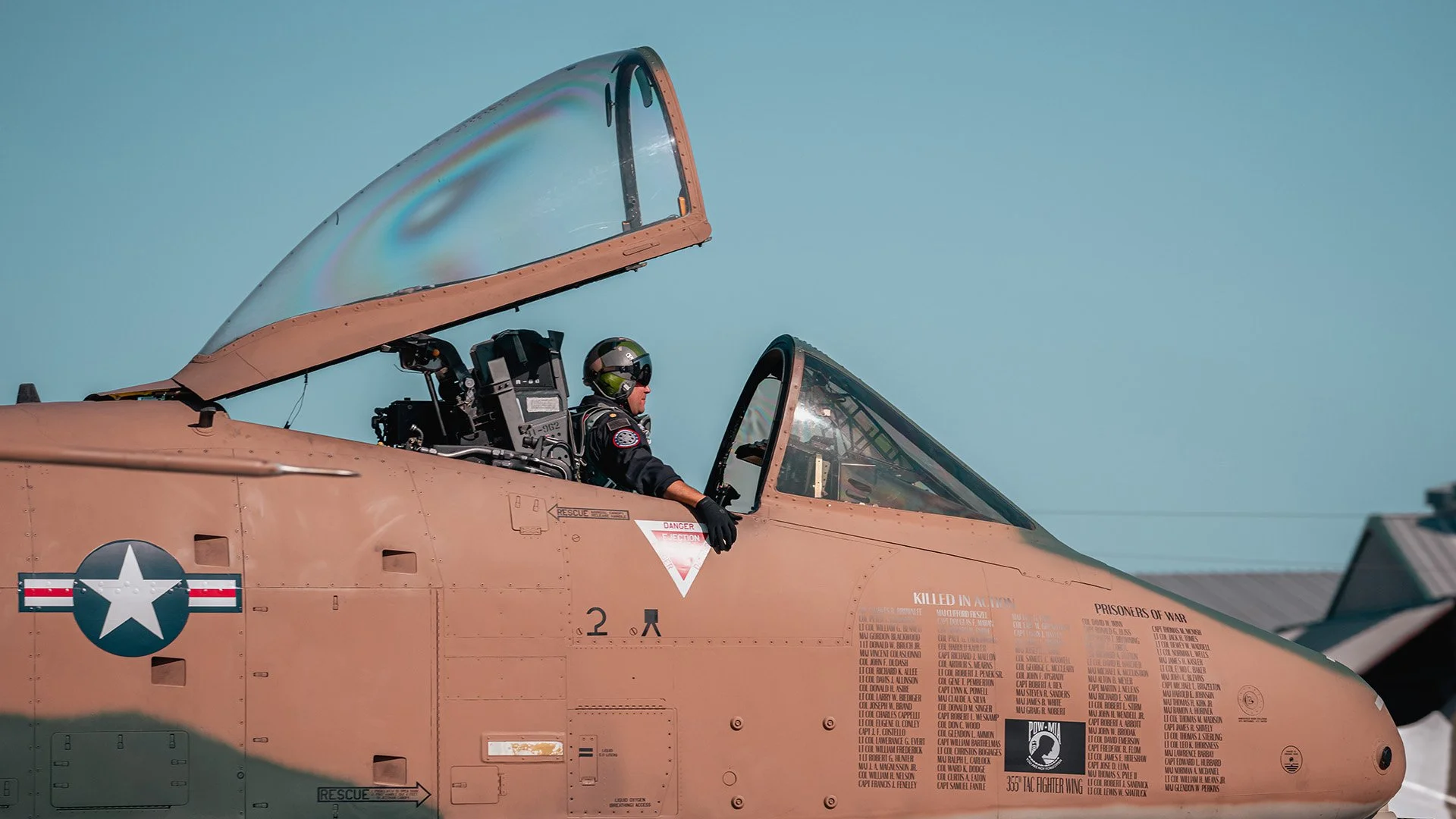 A fighter jet with a pilot in the cockpit, wearing a helmet and gear, under a clear blue sky.