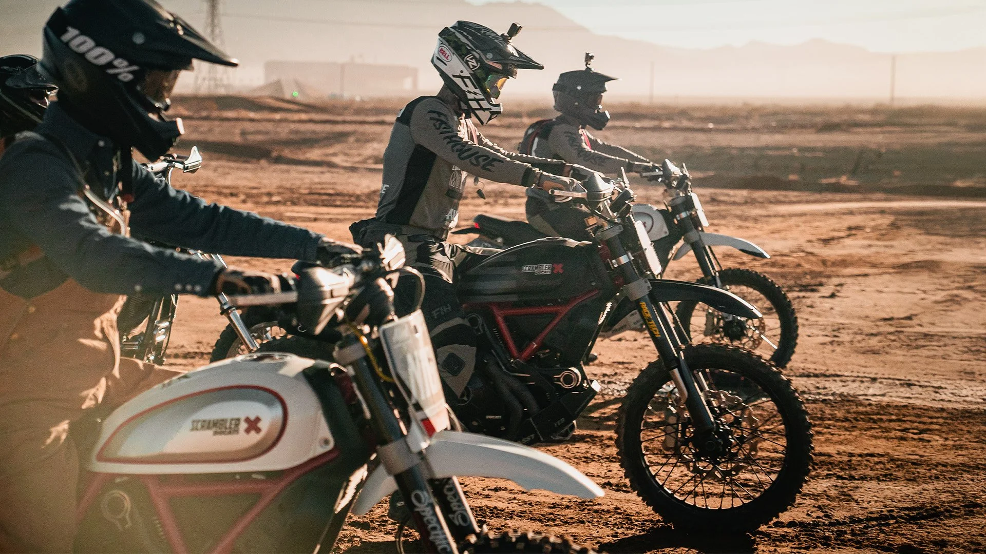 Three motocross riders wearing helmets, goggles, and protective gear, sitting on their dirt bikes on a dusty desert track during sunset.