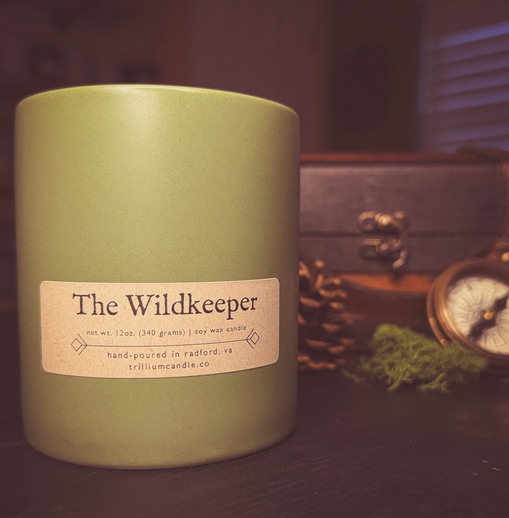 A green soy wax candle with a label that reads 'The Wildkeeper', placed on a dark surface with a small wooden chest and vintage clock in the background.